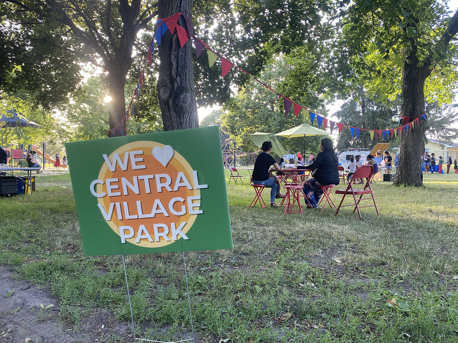 Many people enjoying food and conversations in park, with sign reading "We Love Central Village Park" in foreground