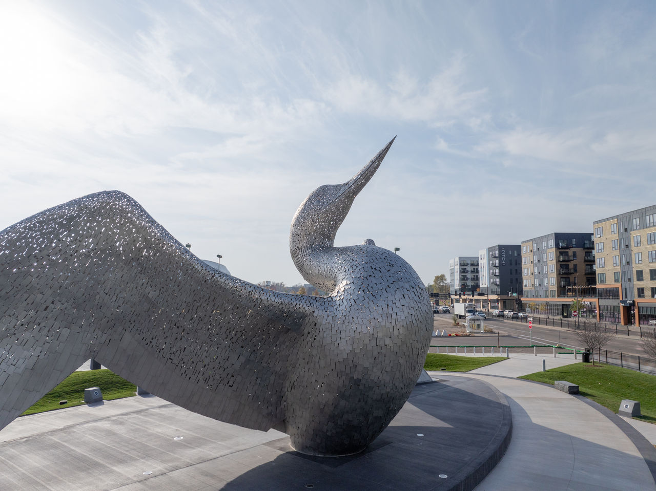 Large steel sculpture of a loon with outspread wings surrounded by green space and apartment buildings in the background.