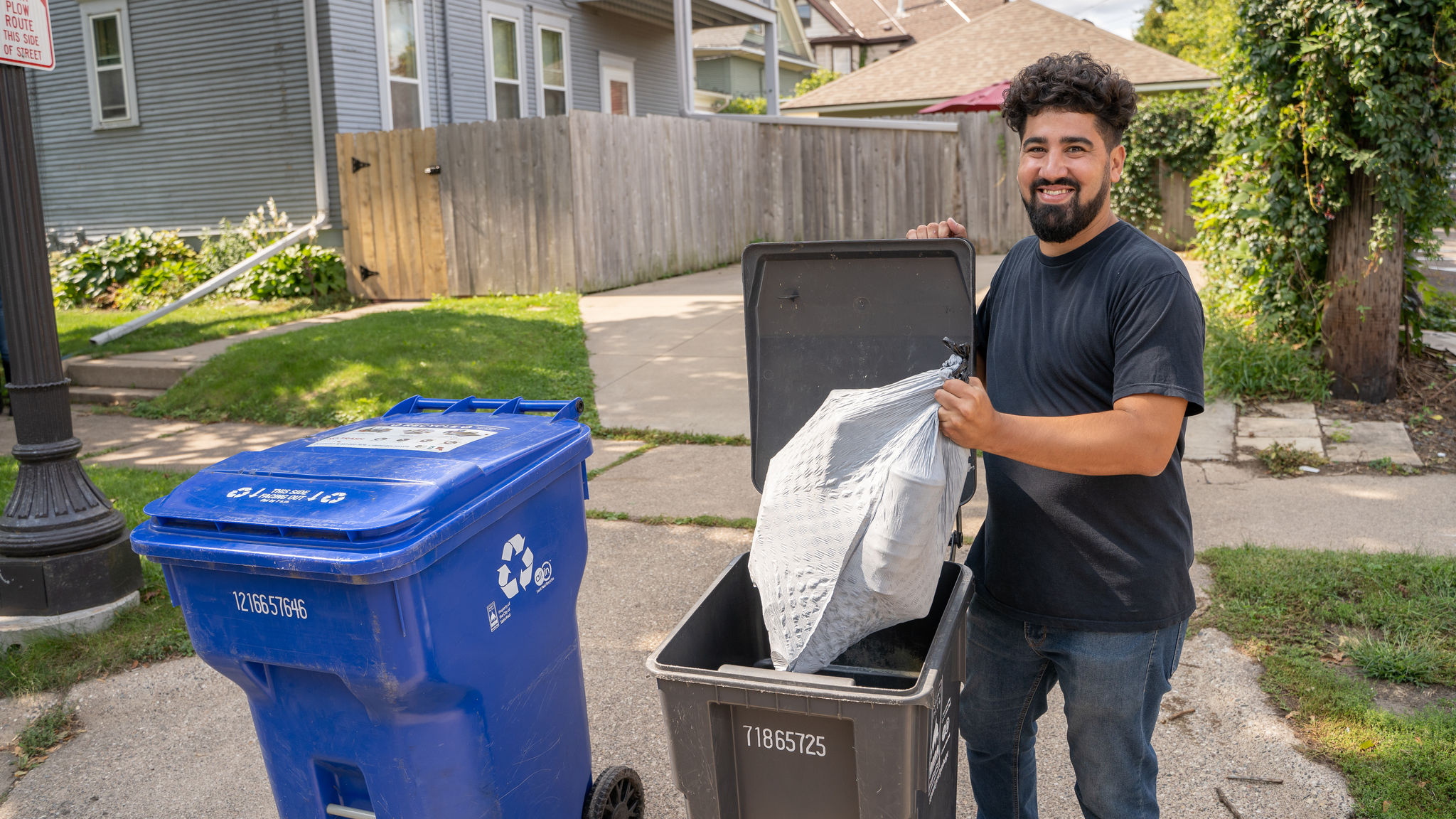 Smiling man placing a plastic bag full of garbage in a black garbage cart.
