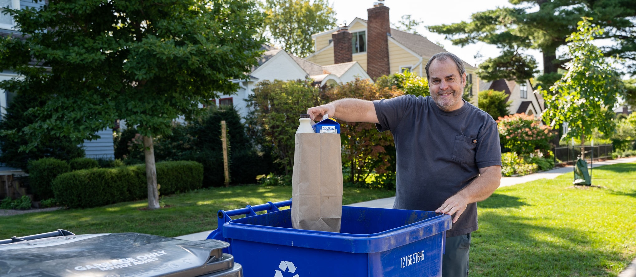 Smiling man placing a paper bag full of recycling in a blue recycling cart.