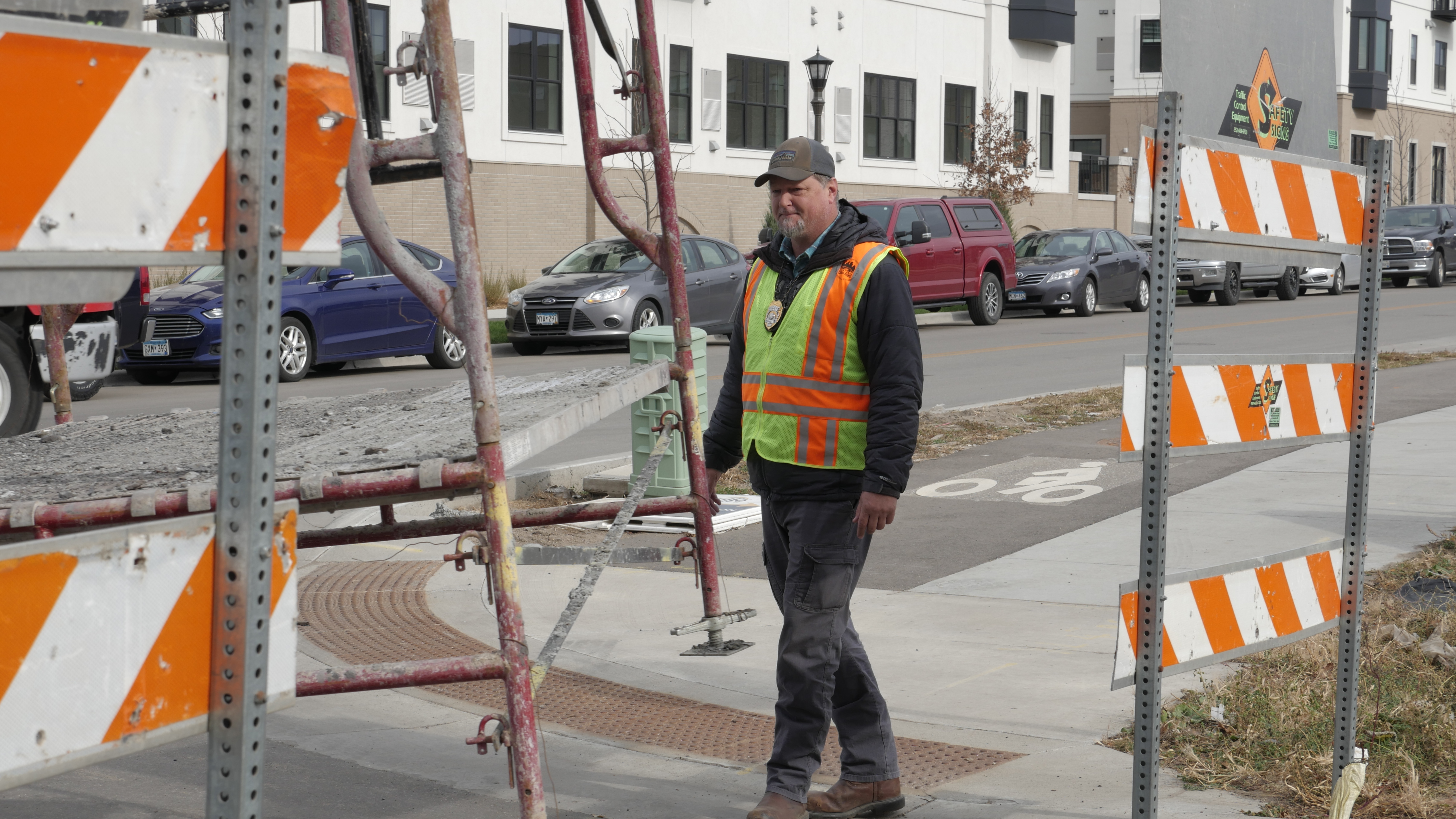 A man wearing a neon safety vest crosses a street behind two orange and white striped construction barriers.