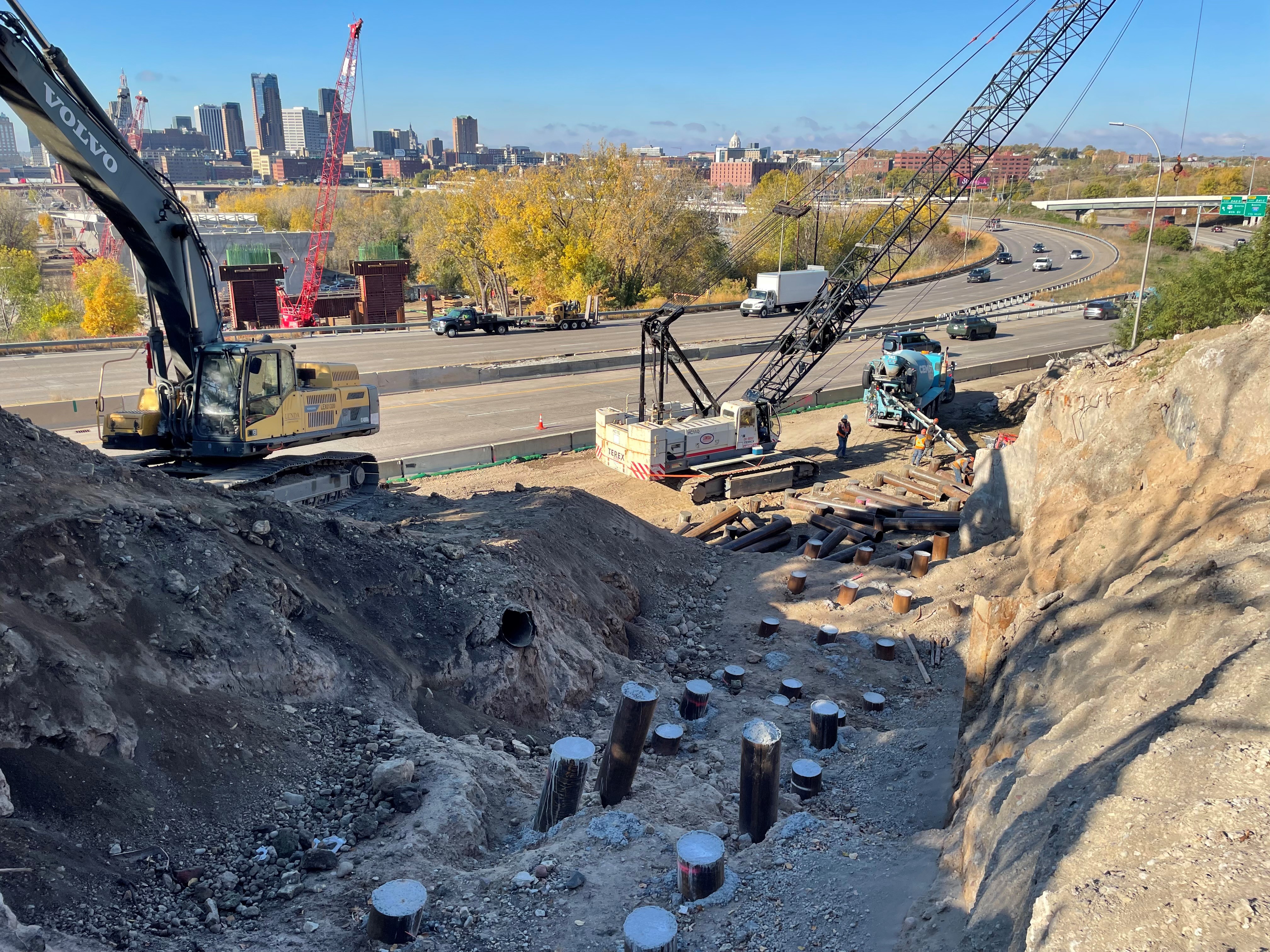 Pipe piles in the ground at the east abutment of the Kellogg-3rd Street bridge with cranes in the background and a view of the downtown Saint Paul skyline.