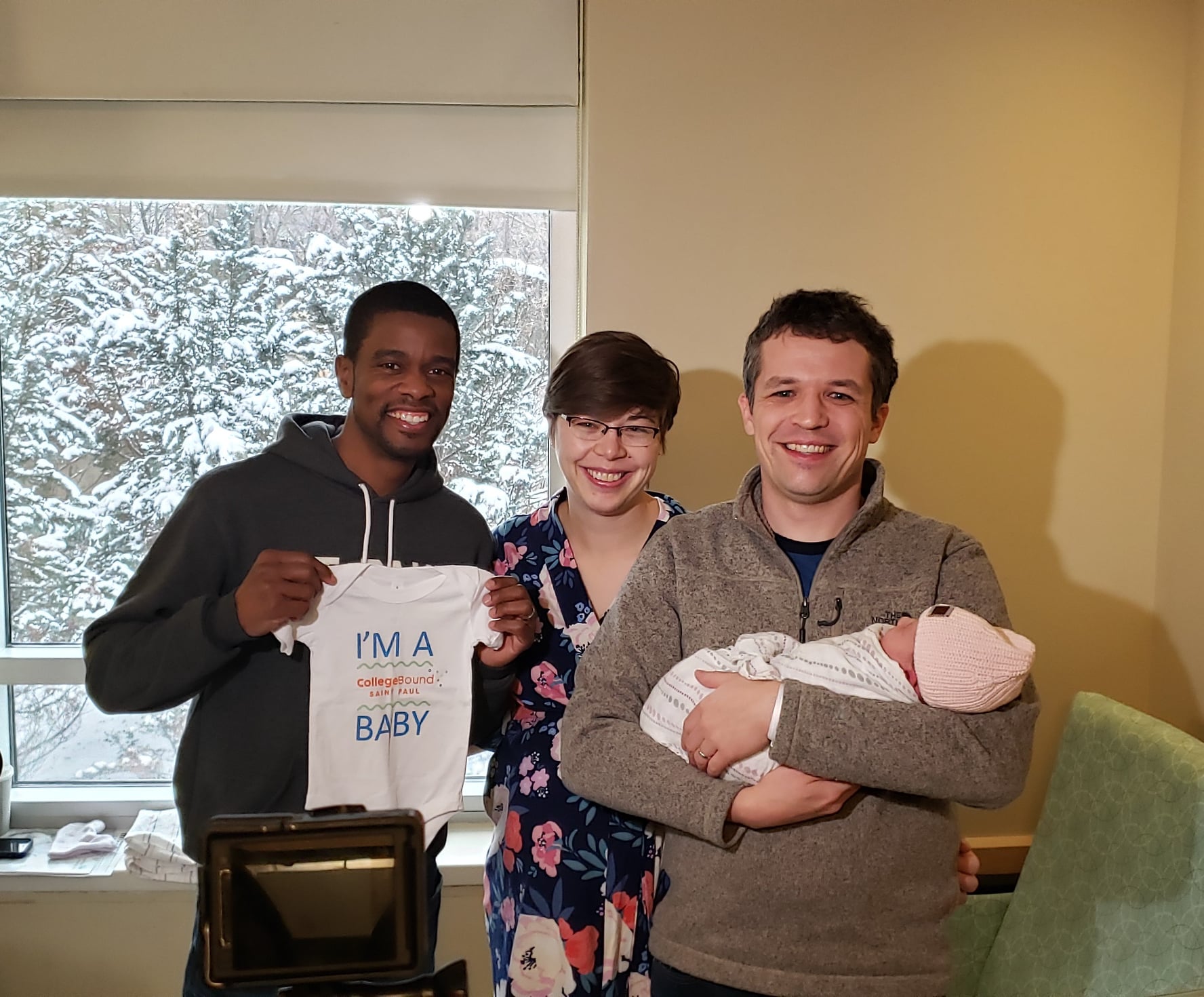 Mayor Carter stands next to a young couple holding a newborn. Mayor Carter holds a onesie that says "I'm a CollegeBound Saint Paul Baby."