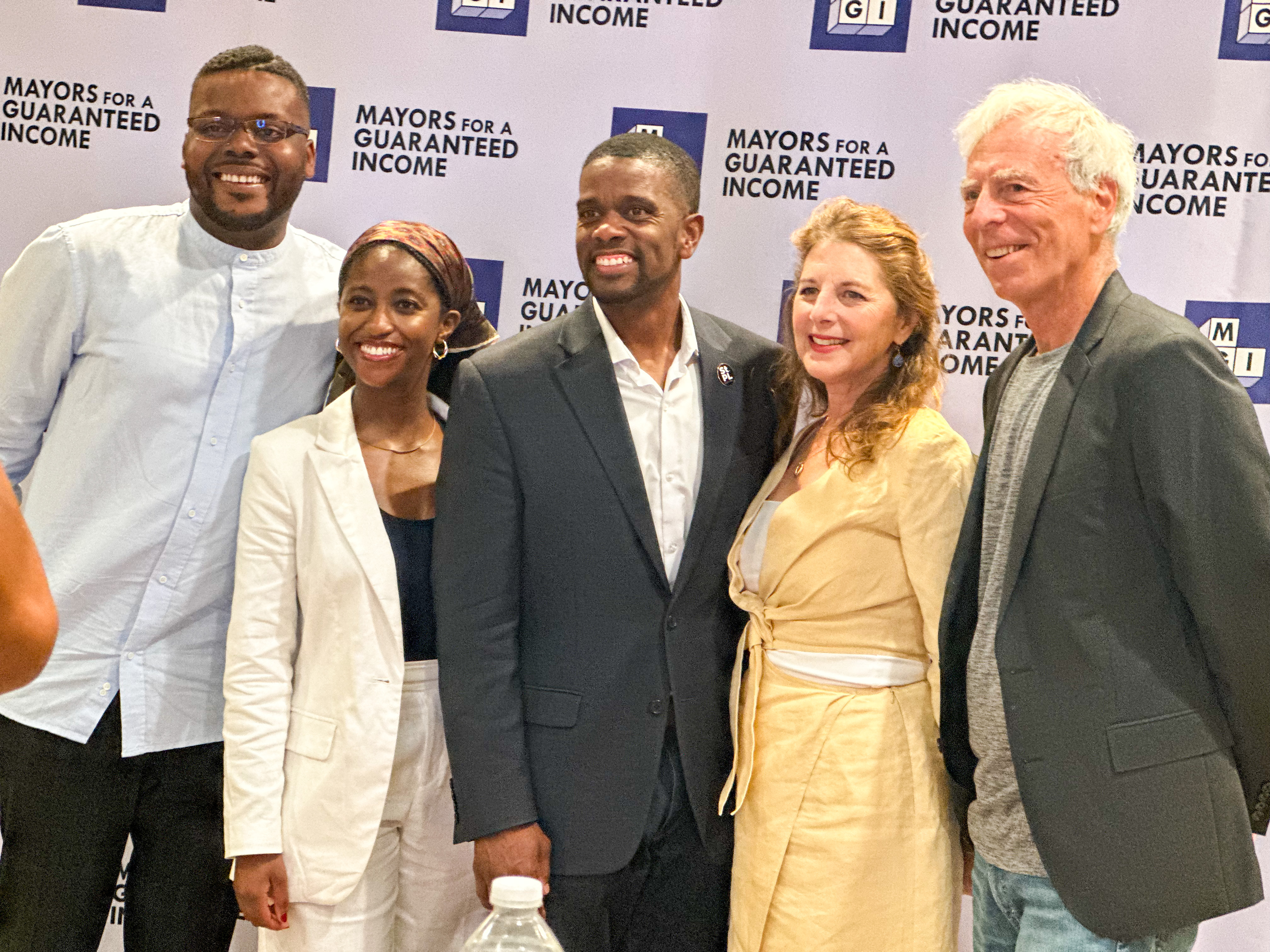 Mayor Carter stands in the middle of a group of professionals in front of a backdrop that says "Mayors for a Guaranteed Income."