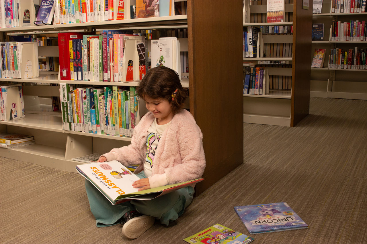 A young school-aged child sits on the floor next to library shelves of children's books. She has two books sitting beside her, and is reading a third.