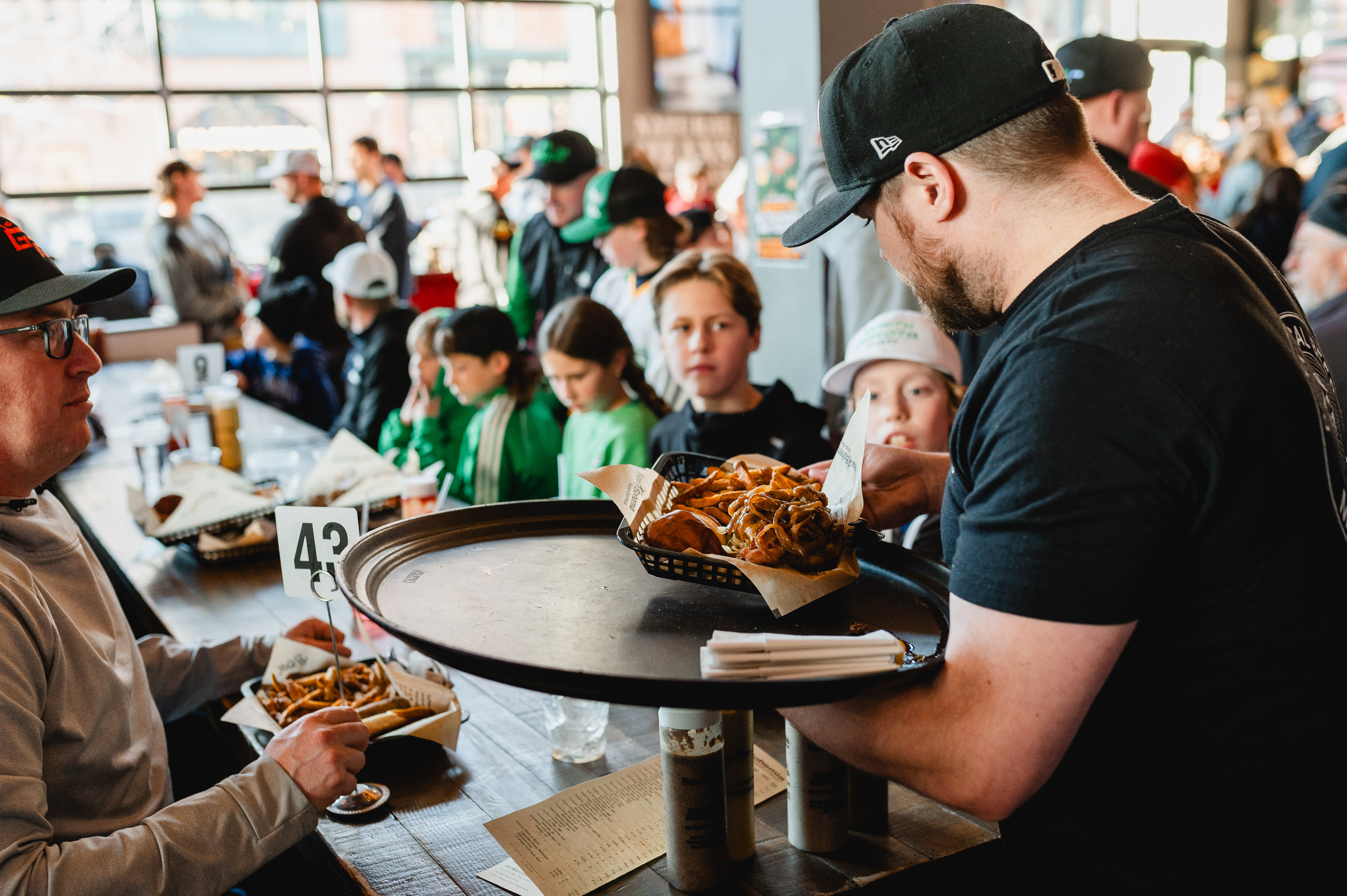 A server brings burgers and fries to a family in sports memorabilia. They're seated in a large open restaurant with garage door windows.