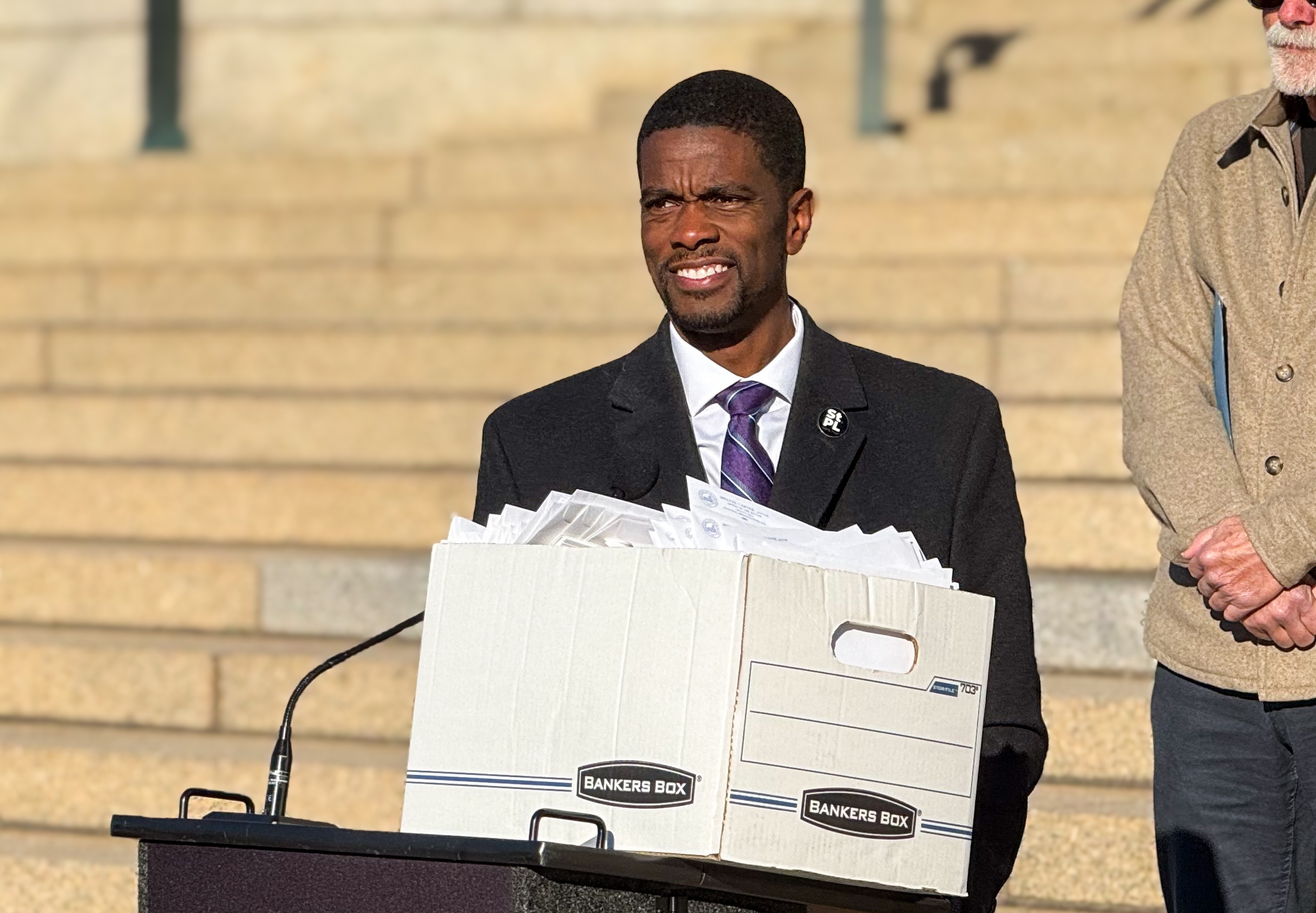 Mayor Carter holds an overflowing box of paperwork while speaking at a podium outside in the sunshine.