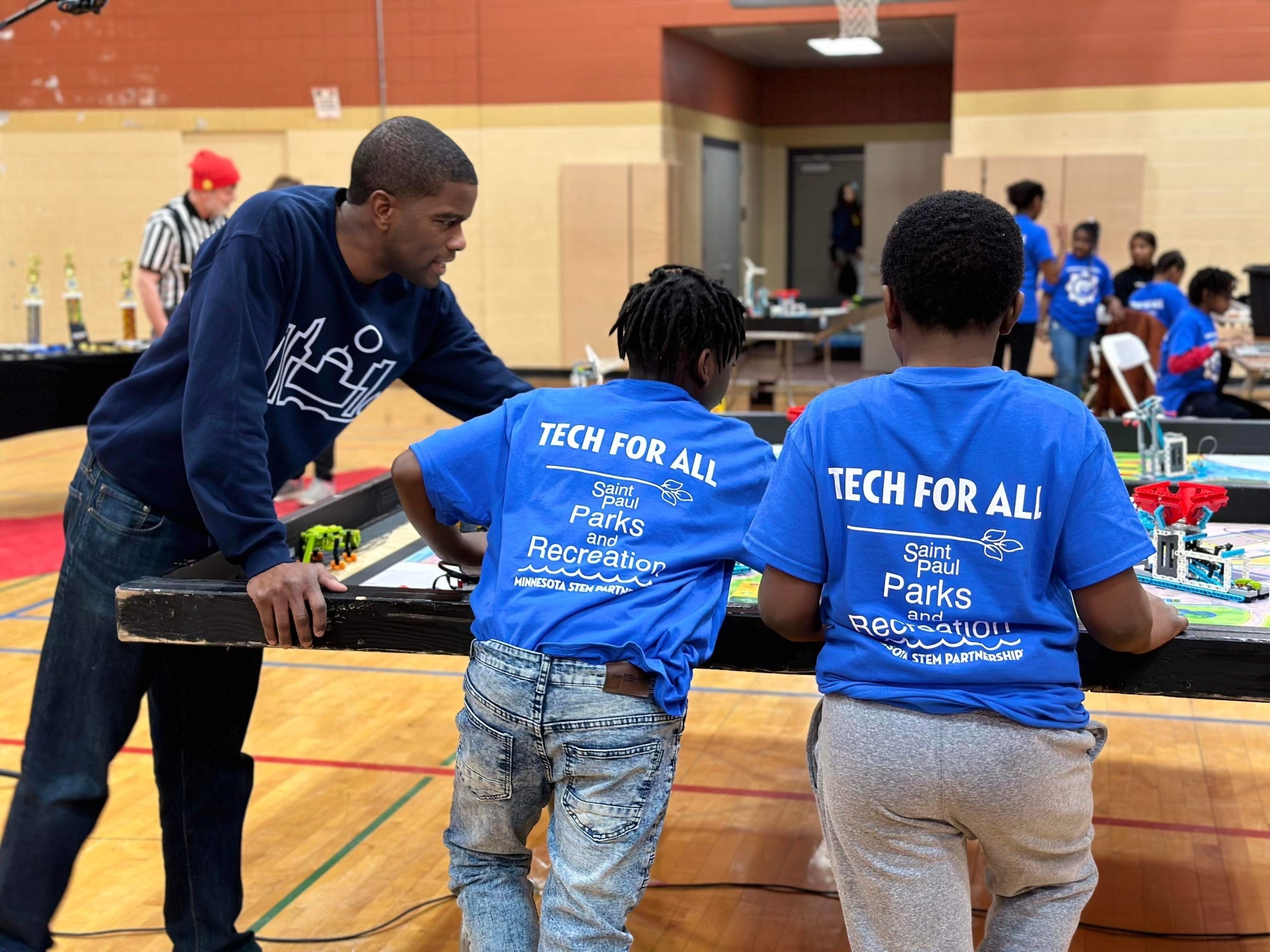 Mayor Carter leans over a table in a gym with two young children in Tech For All t-shirts. They're looking at a series of lego robotics.
