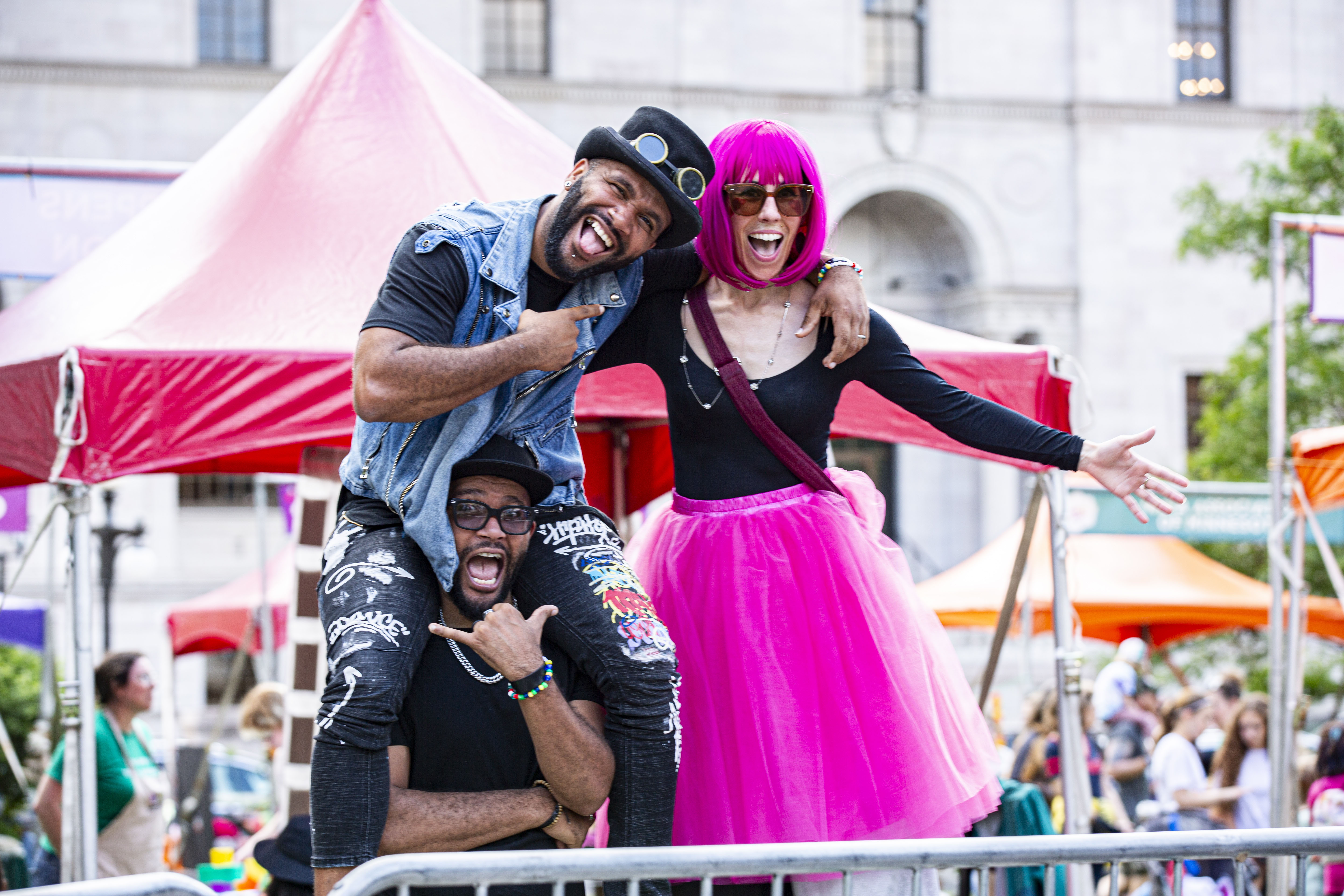 Three people in fun dress stand downtown in front of a crowd and several tents. They smile and pose for the camera.