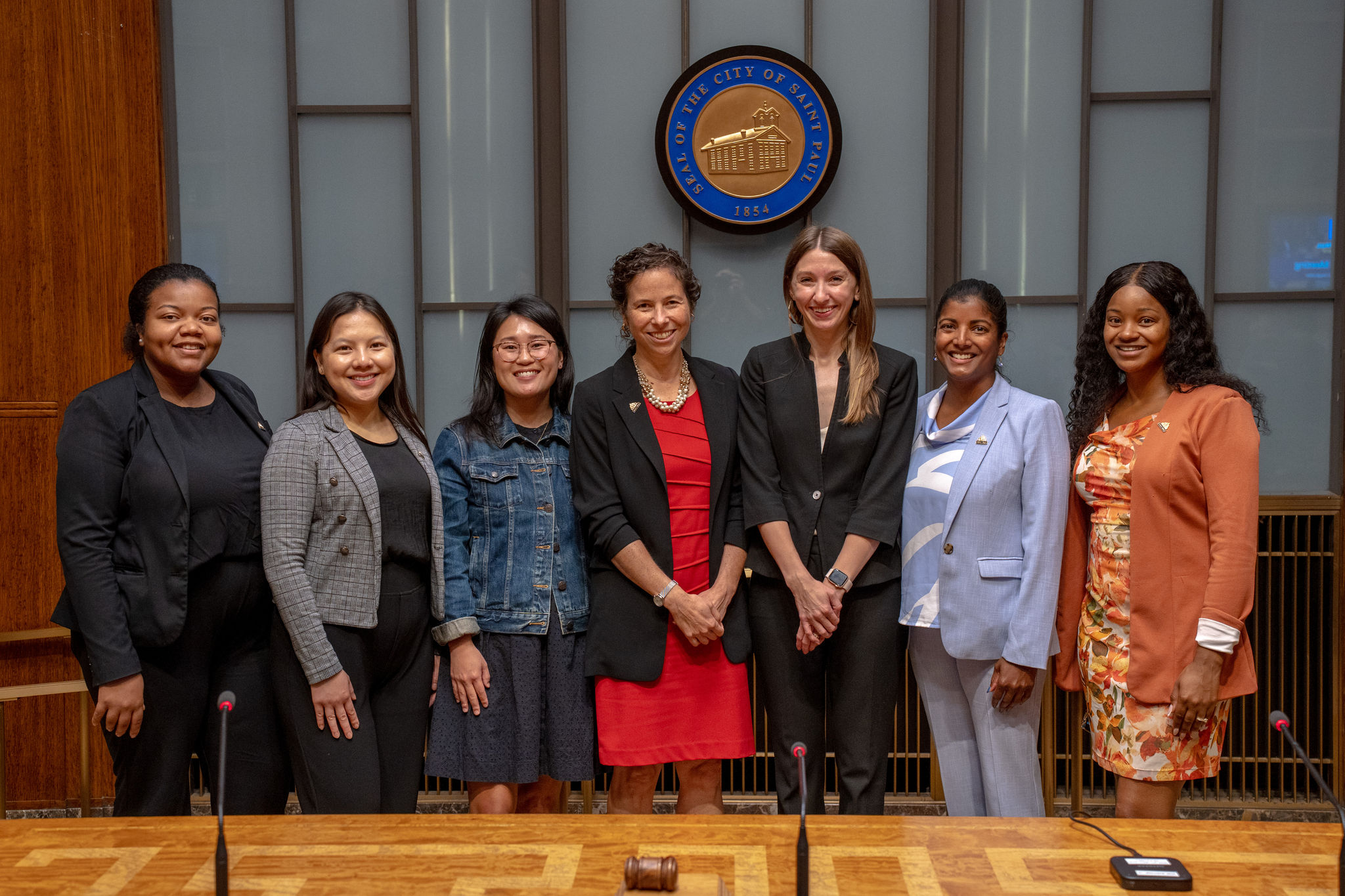 Seven diverse women stand beneath the official seal of the City of Saint Paul, in front of a polished table with microphones on it.