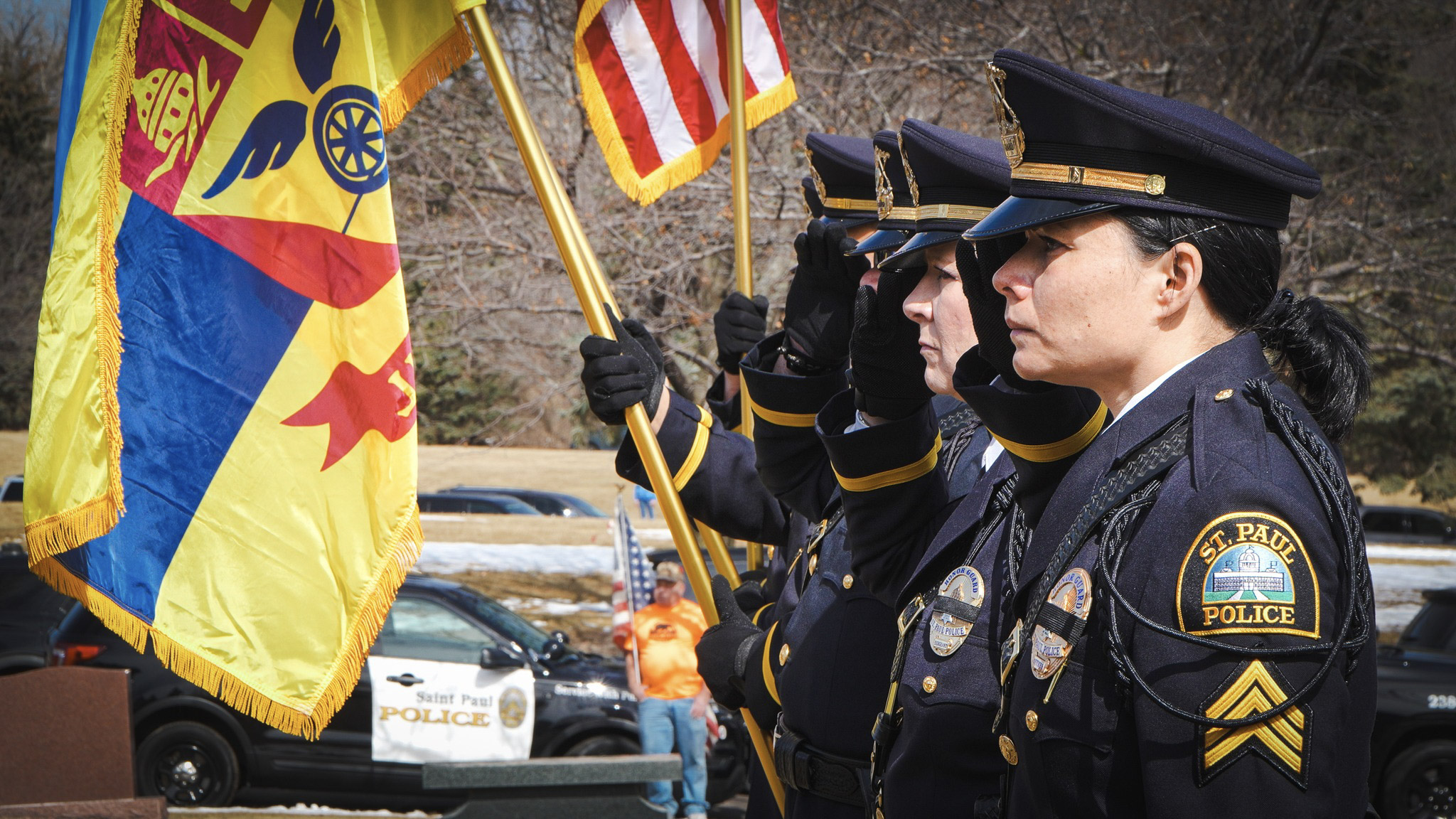 Women from SPPD stand in uniform saluting and holding the flags of Saint Paul and the United States.