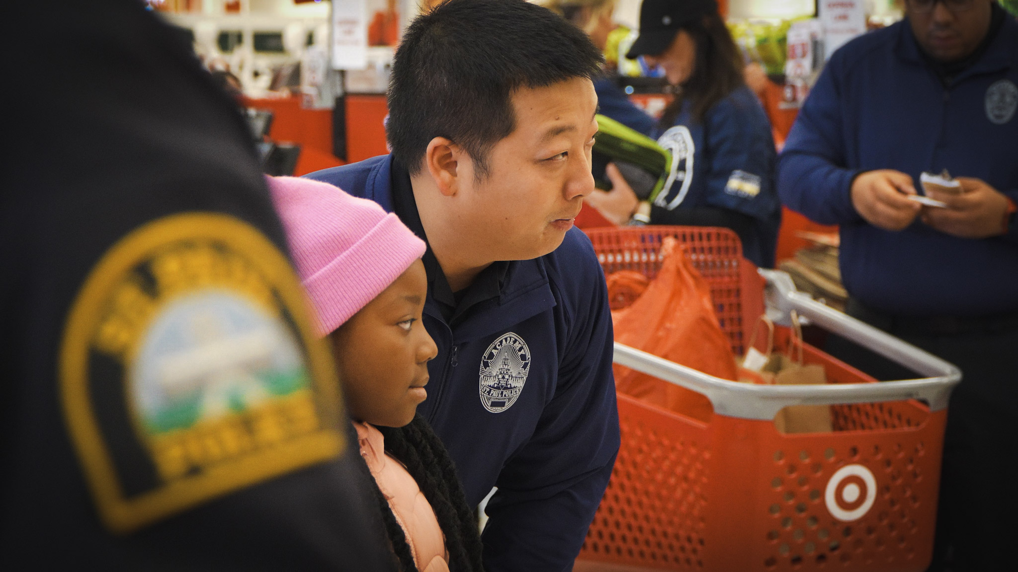 A police officer in a casual jacket talks with a school-age child at Target. Other shoppers walk by and an additional officer looks on.