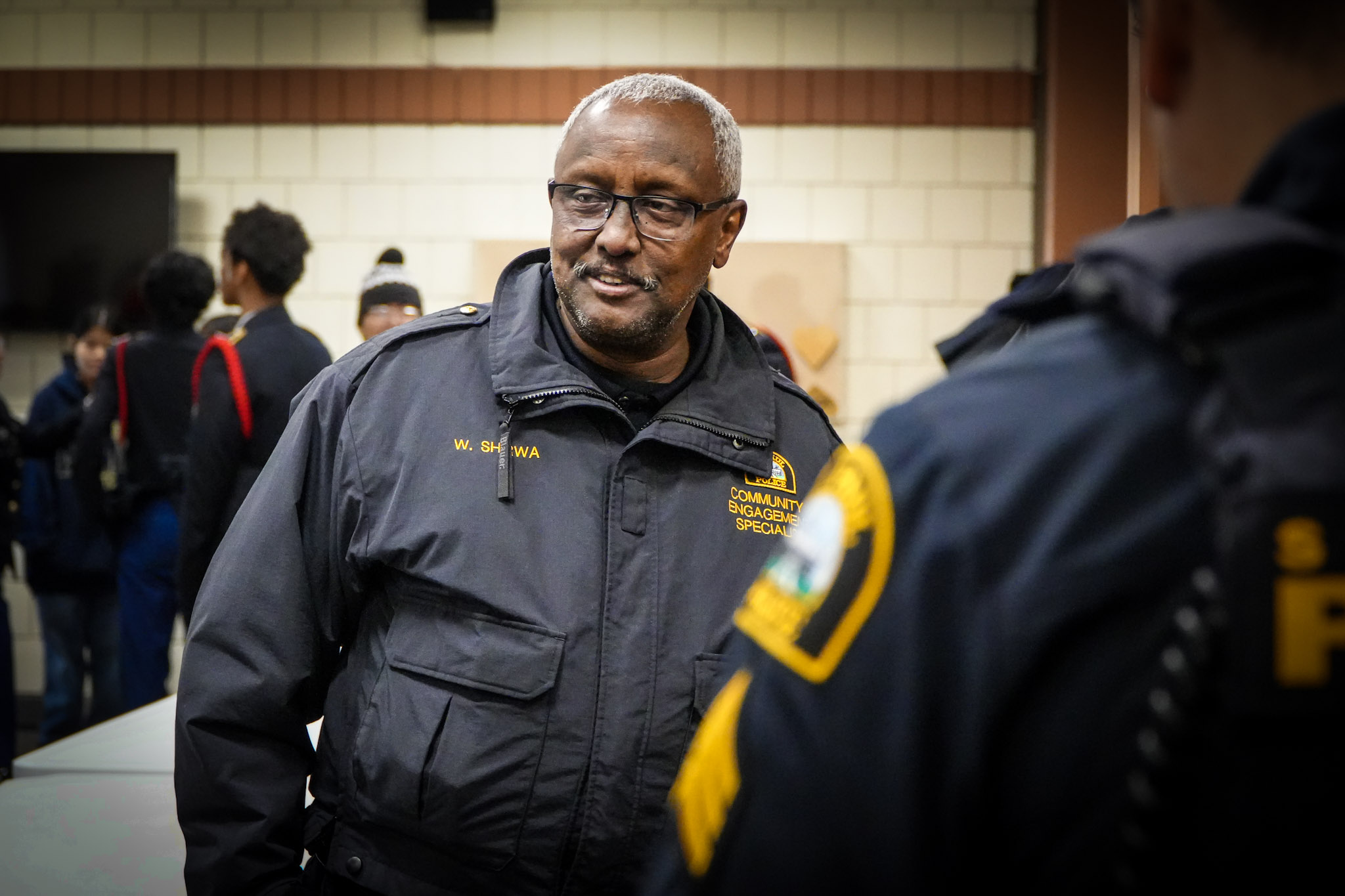 An older black man in an SPPD community liaison officer windbreaker. He talks with officers and young people in a school gym.