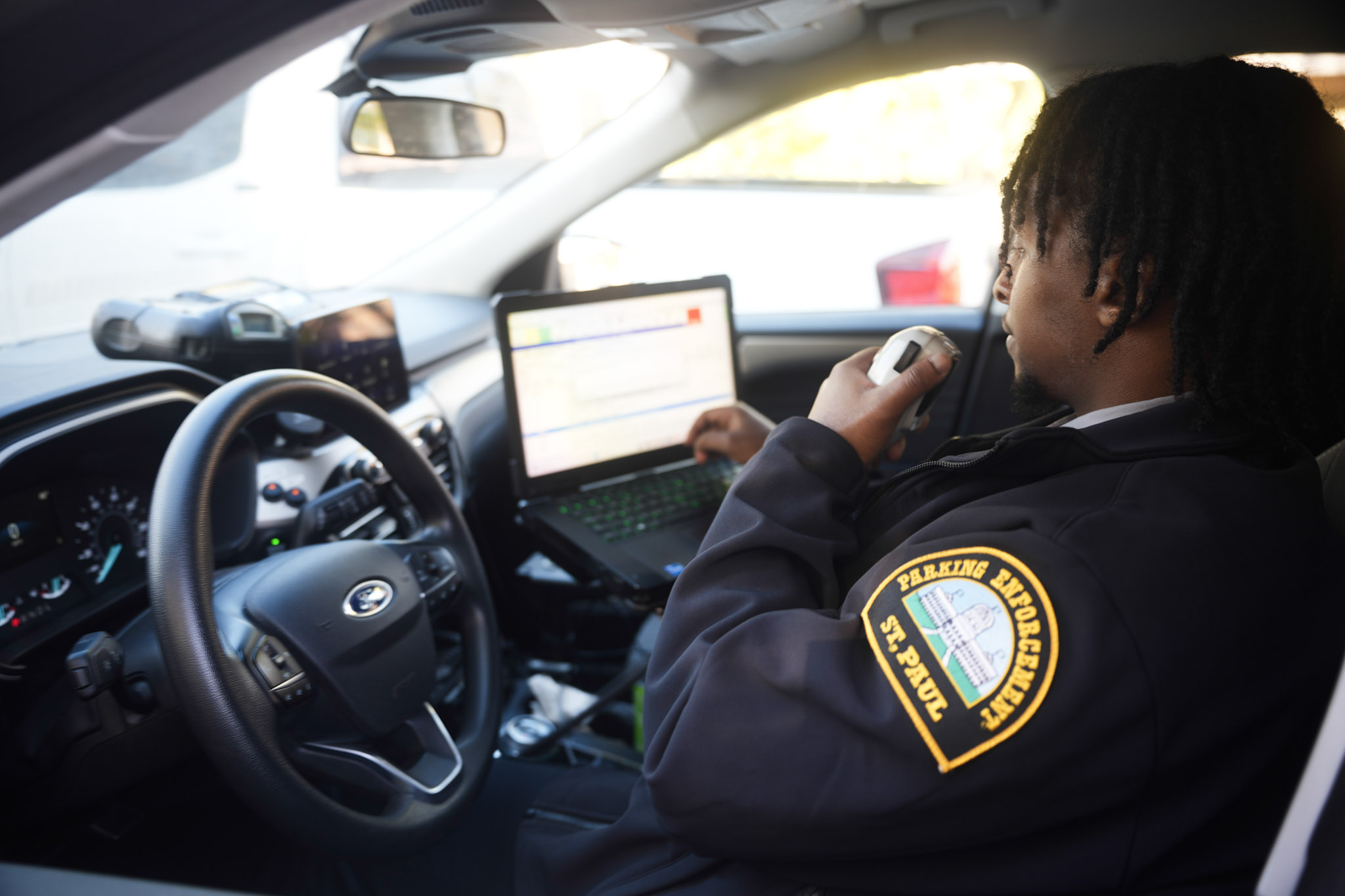 A young black man in an SPPD Parking Enforcement Uniform radios a colleague while looking something up on a laptop while parked in his car.