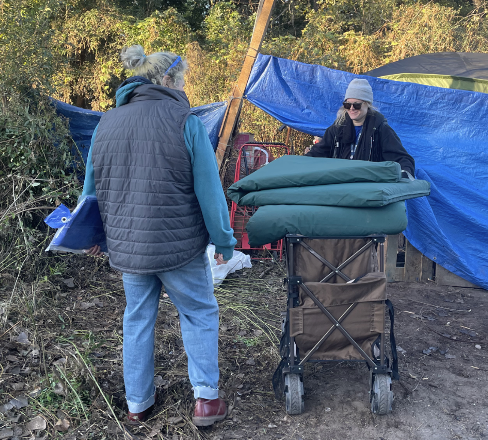 Two women talk outside a makeshift campsite under blue tarps. They are bringing supplies.