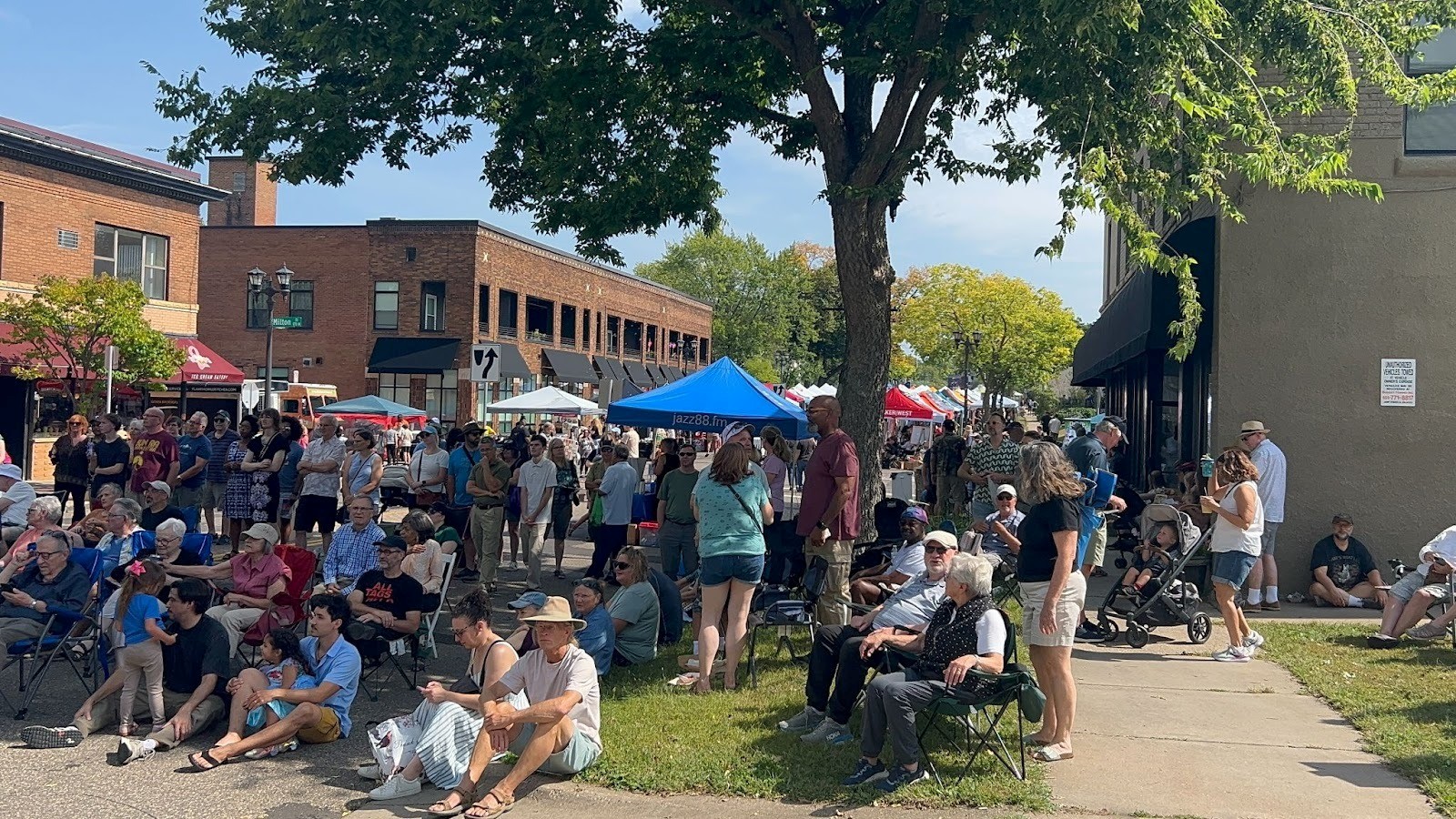 Attendees gather in lawn chairs under shade trees at the Selby Avenue Jazz Festival, with vendor tents lining the street behind them.