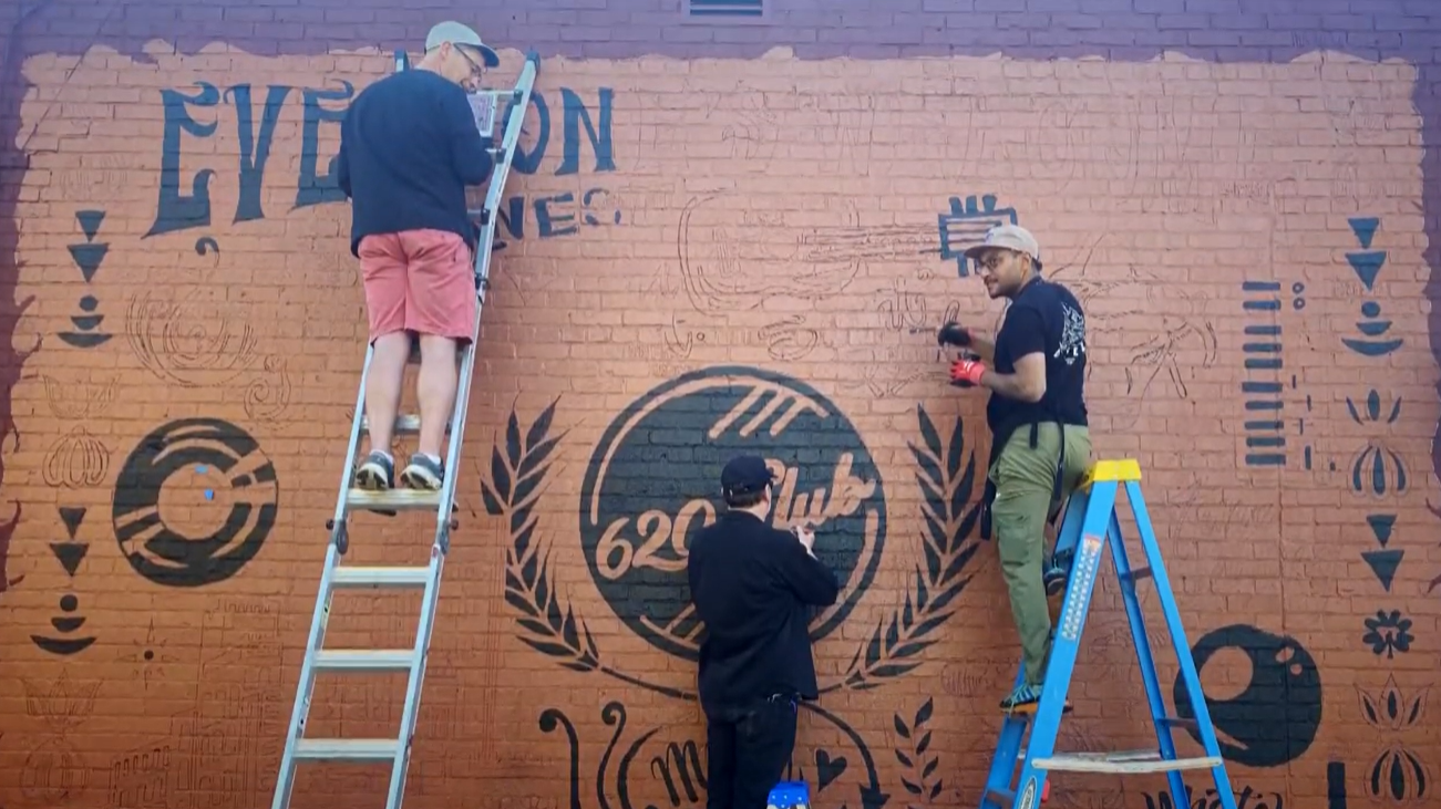Three artists on ladders paint a mural featuring a 620 Club emblem on a brick wall.