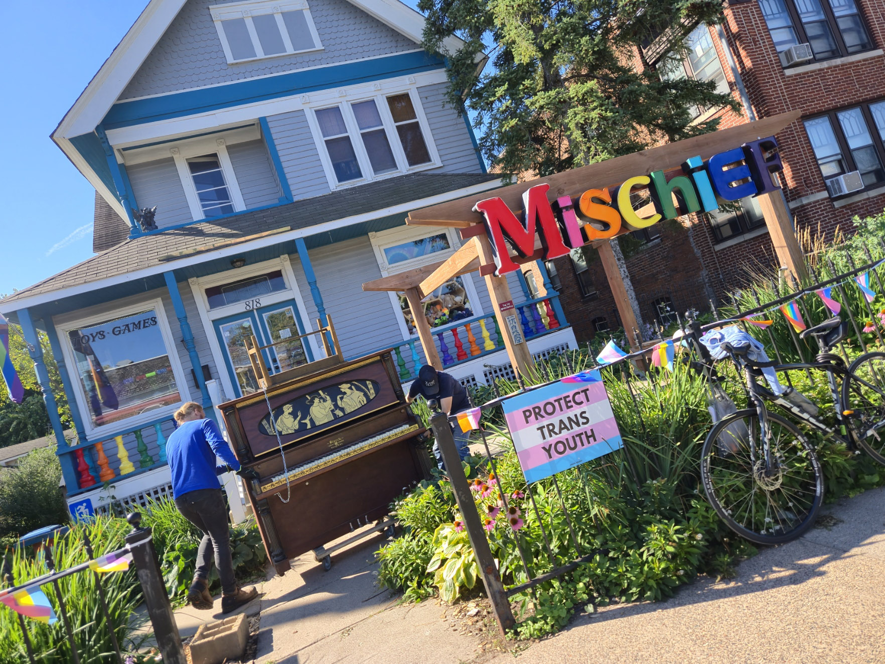 Two people move a piano outside Mischief toy store, a colorful house decorated with flags