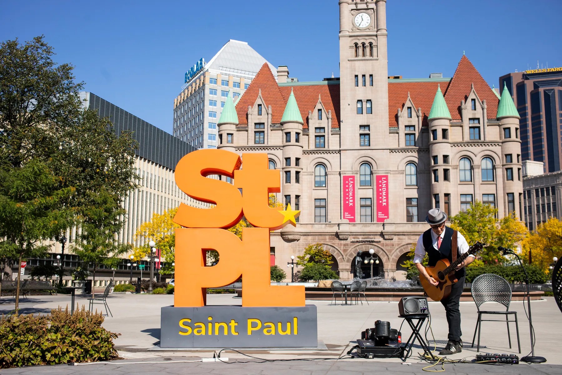 A guitarist performs at a lunchtime concert in Rice Park, with the orange "StPL Saint Paul" sculpture and the Landmark Center behind.