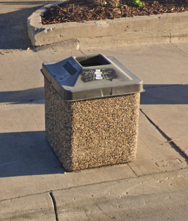 A concrete trash bin on a sidewalk beside a planter.