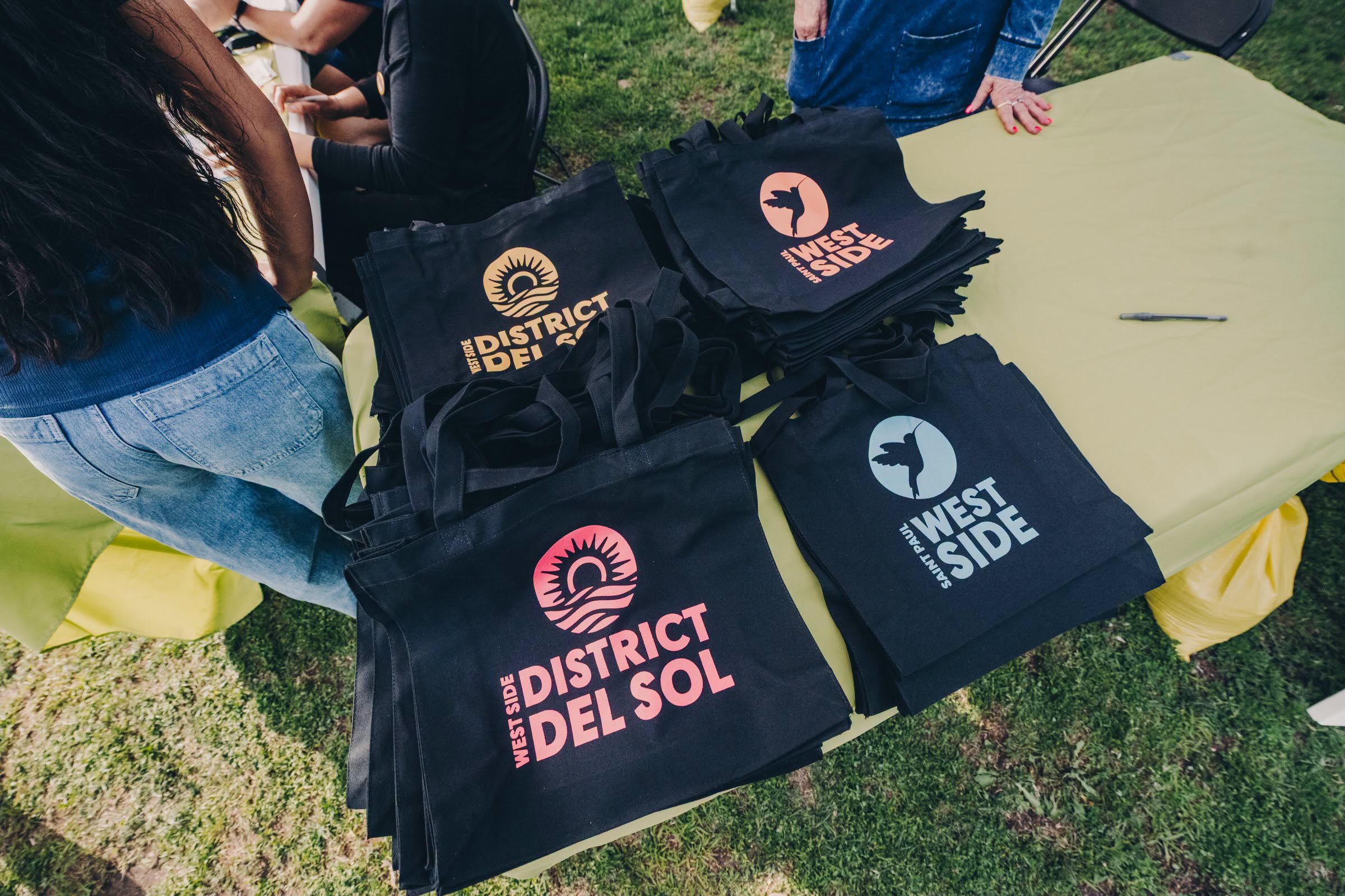 Black tote bags printed with "West Side District del Sol" and "Saint Paul West Side" branding on an event table.