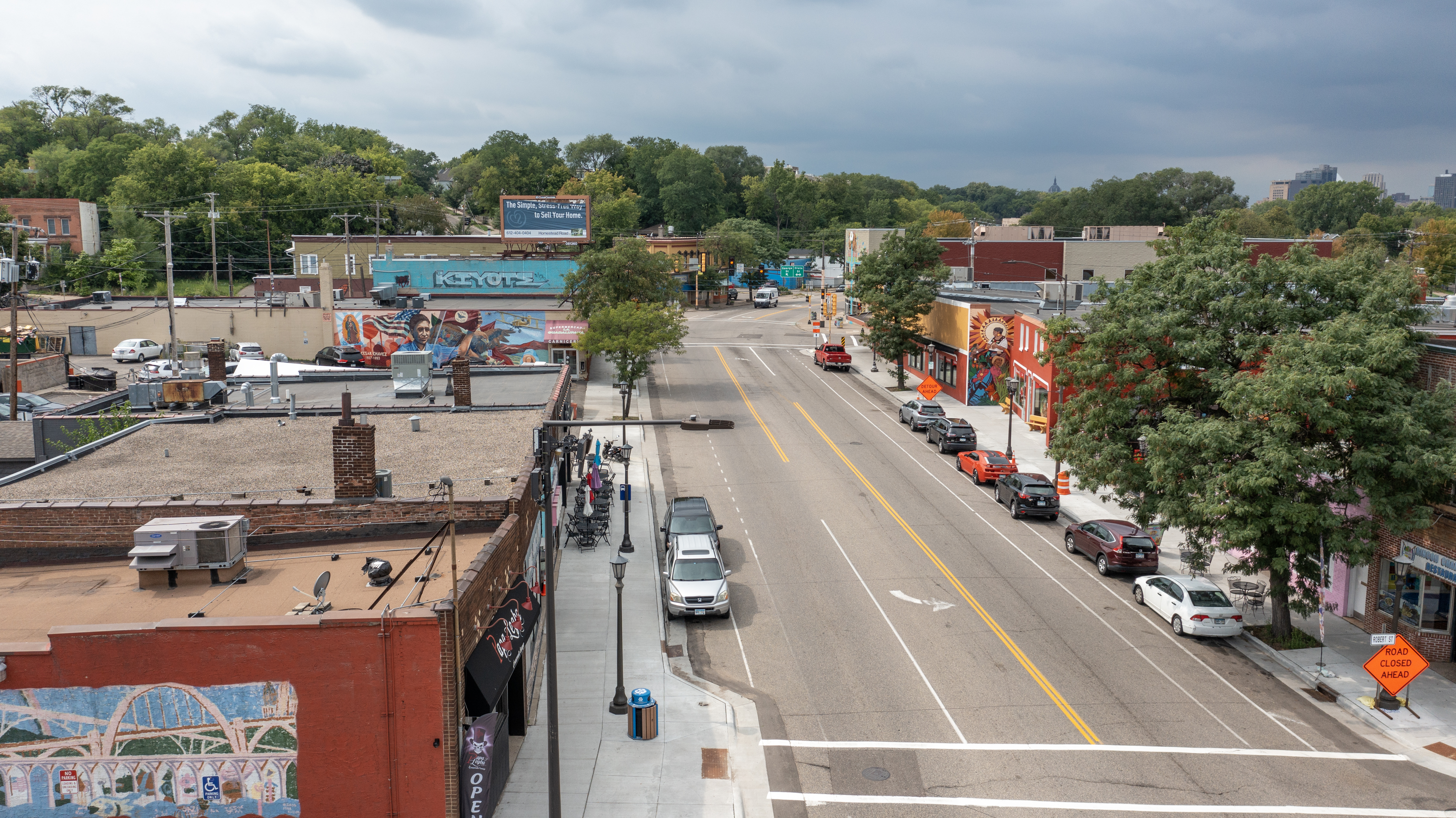 Aerial view of César Chávez Street on the West Side, with brick storefronts, murals, and the Saint Paul skyline in the distance.