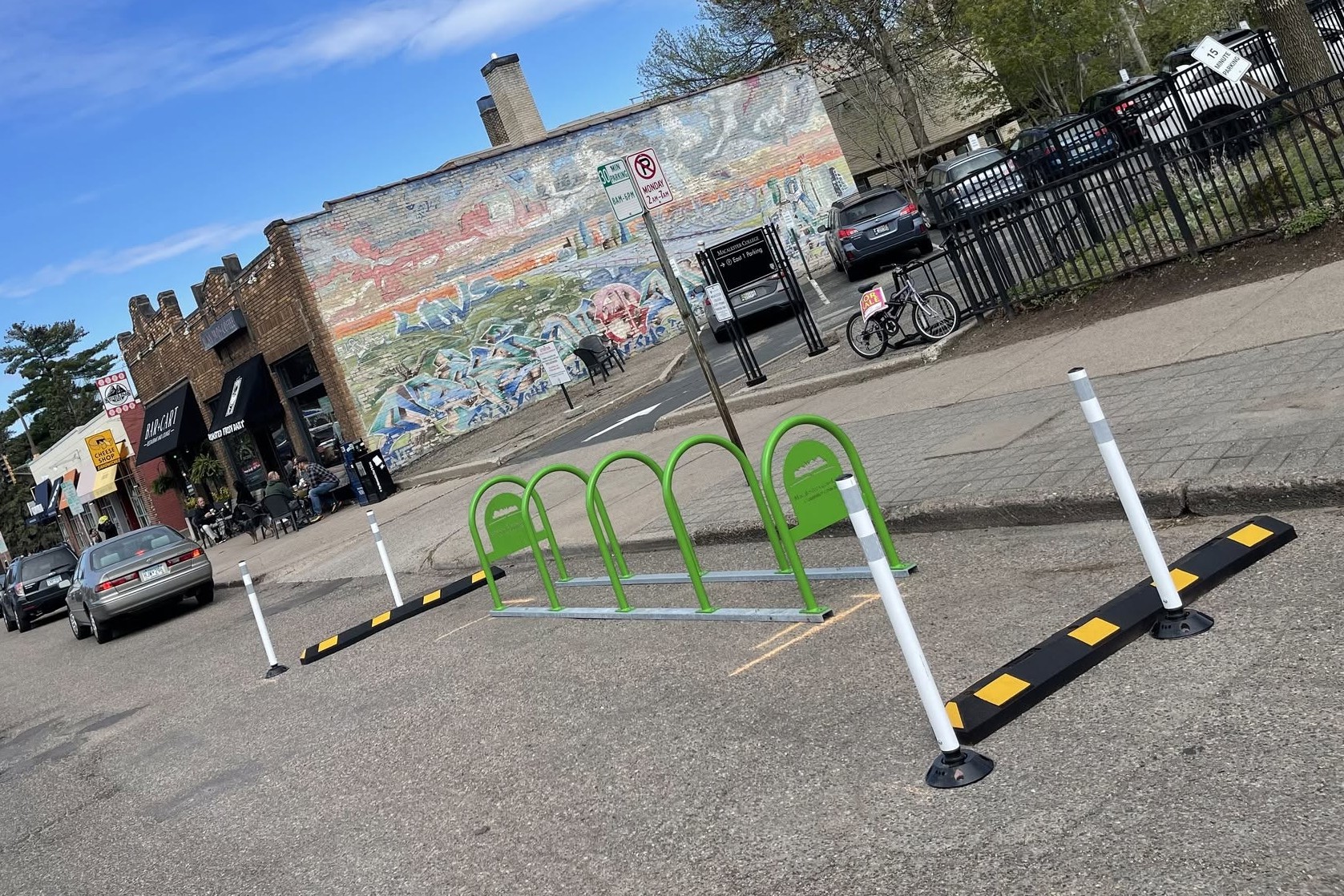 A row of five green Macalester-Groveland Community Council bike racks installed in a converted on-street parking space on Grand Avenue, with a mural and storefronts across the street.