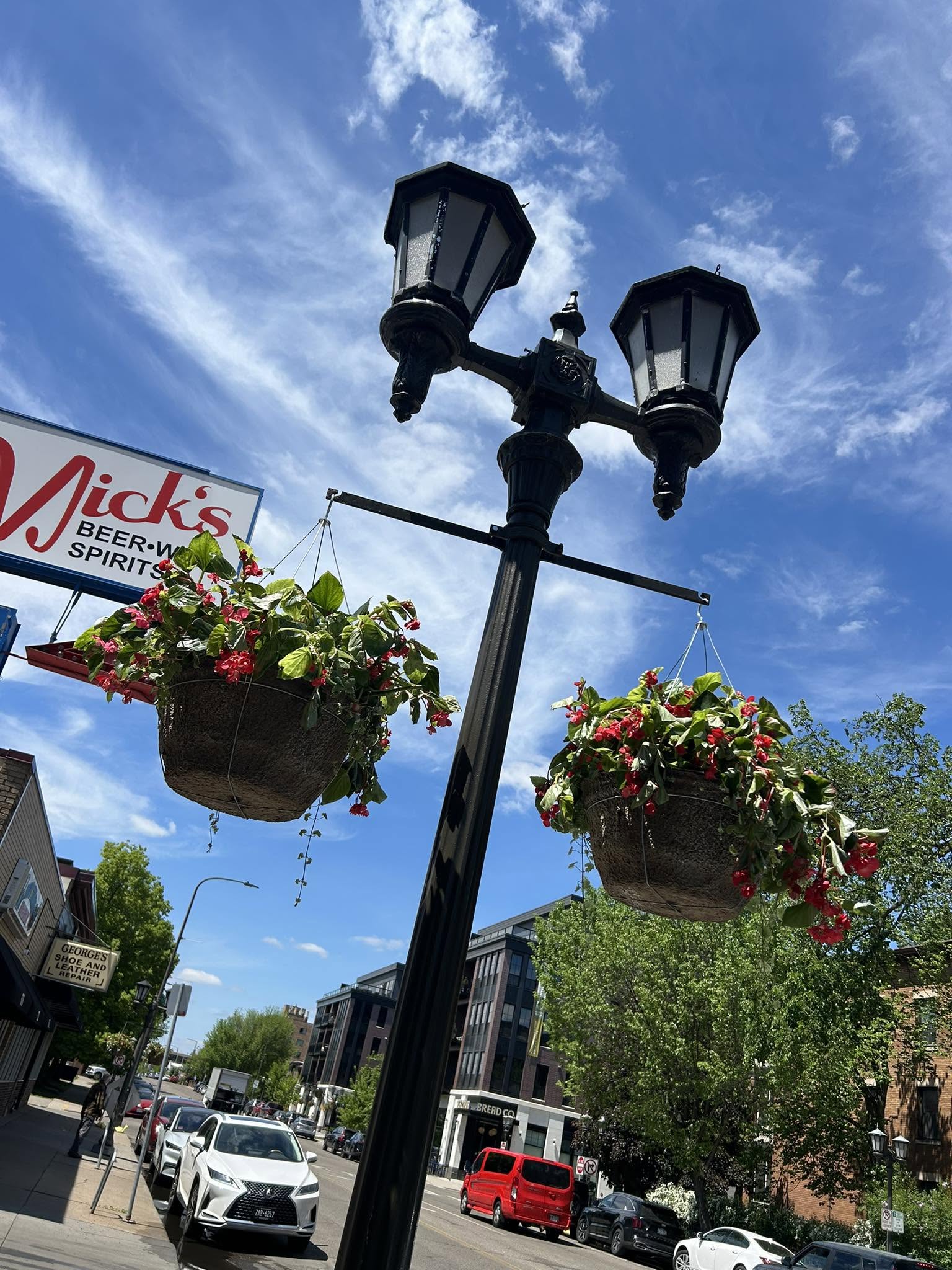 Hanging baskets of red begonias on an ornate black streetlamp along Grand Avenue, with the Nick's Beer, Wine & Spirits sign in the background.