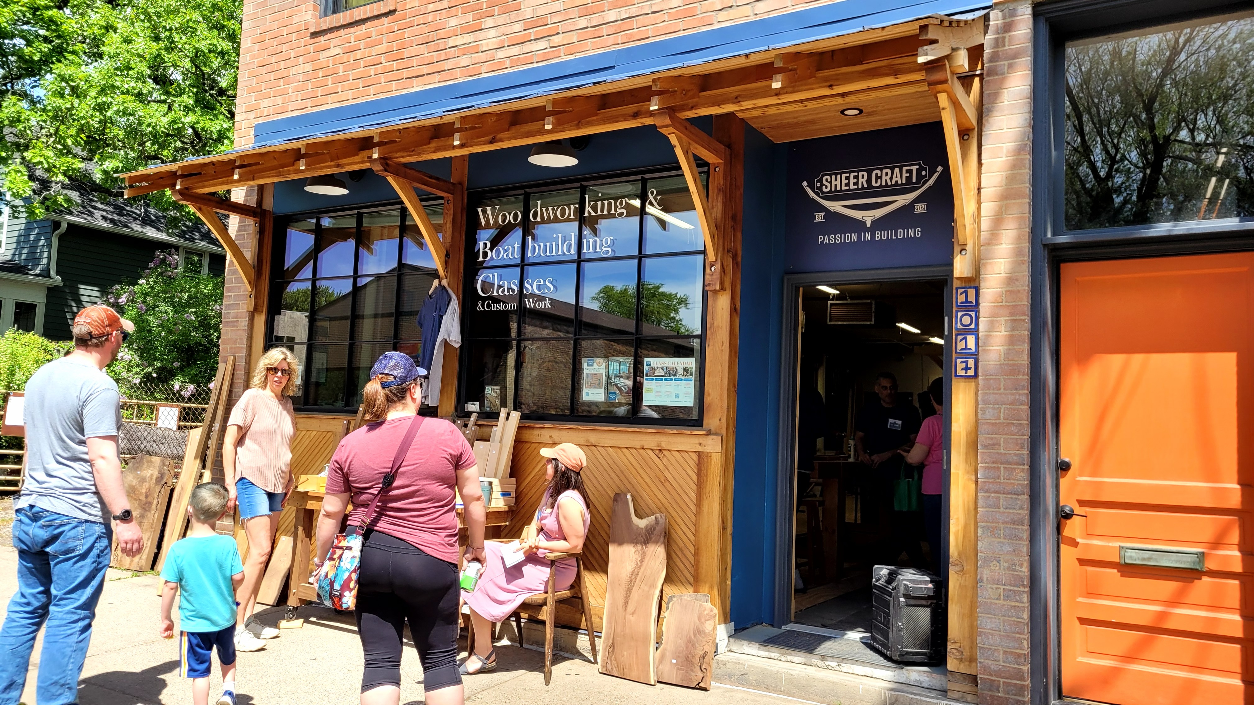 Sheer Craft woodworking shop storefront with visitors browsing outside during an open-shop event, showing signage for woodworking and boat-building classes.
