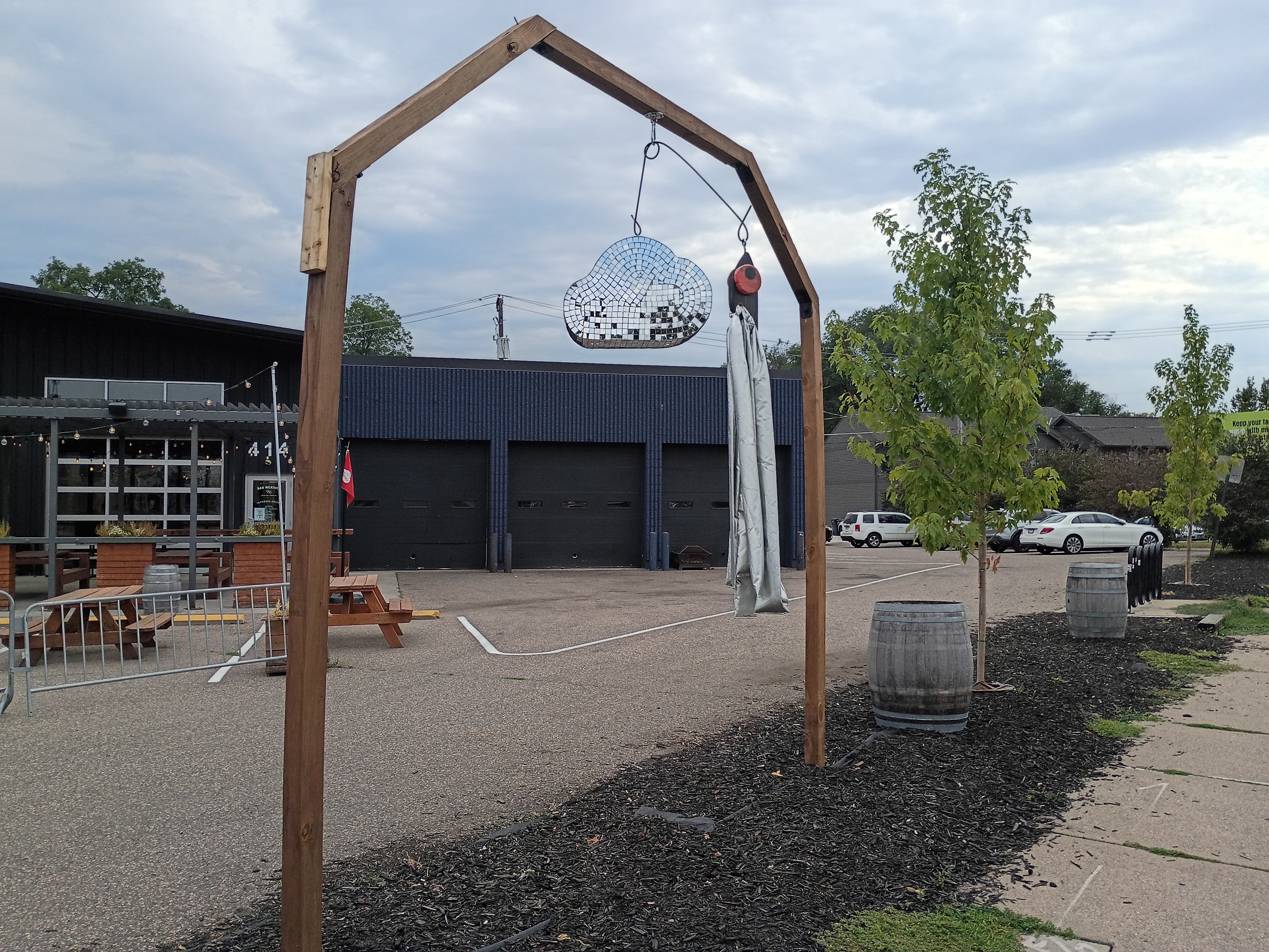 Wooden peaked-arch entryway with a silver mosaic cloud sculpture in front of Bad Weather Brewing at 414 W. 7th Street, with the brewery's patio and wine barrel planters visible.
