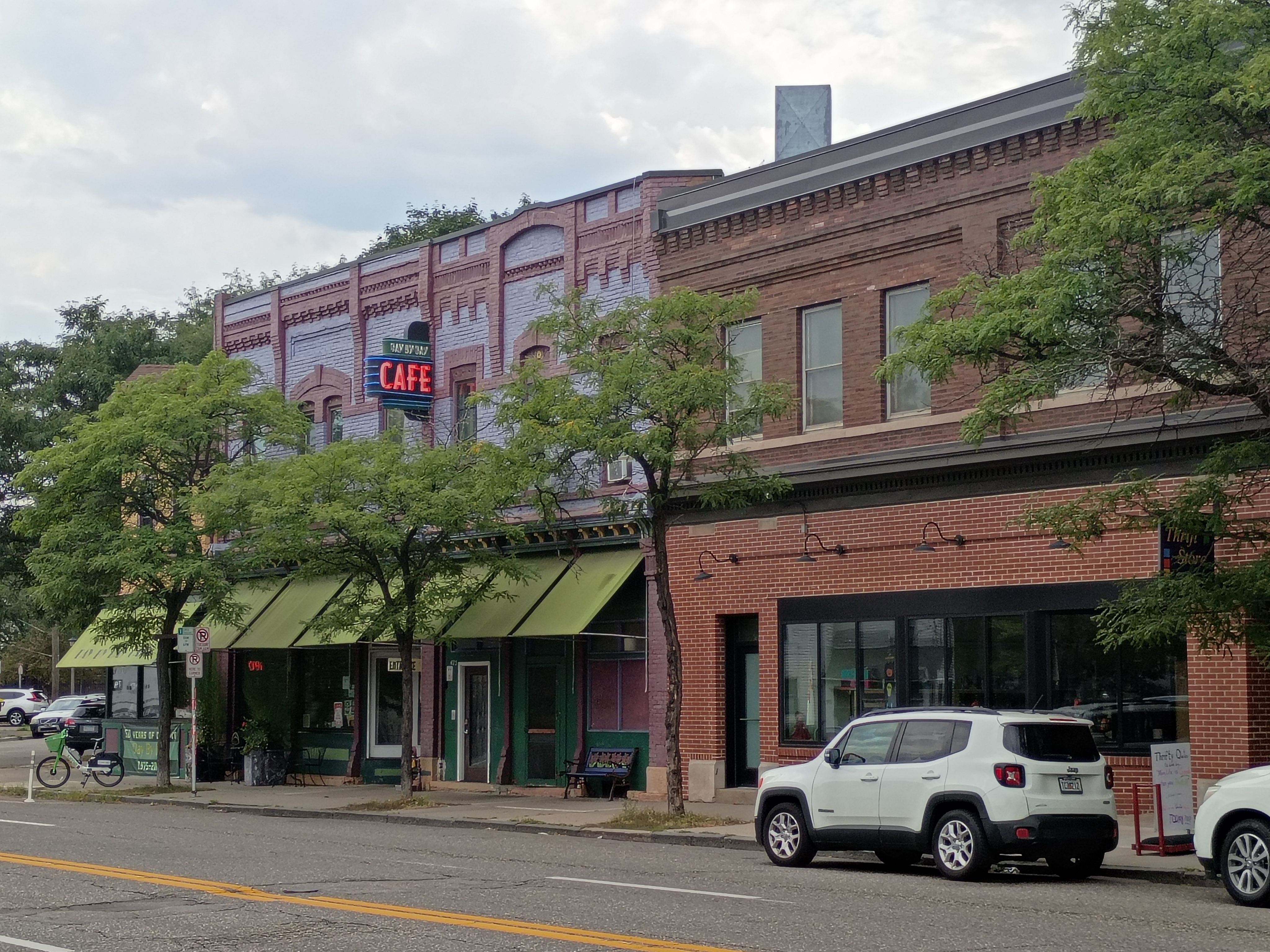 Row of West 7th Street storefronts including Day by Day Cafe in a lavender-painted historic brick building with green awnings, next to a red brick building housing a Thrift Store.