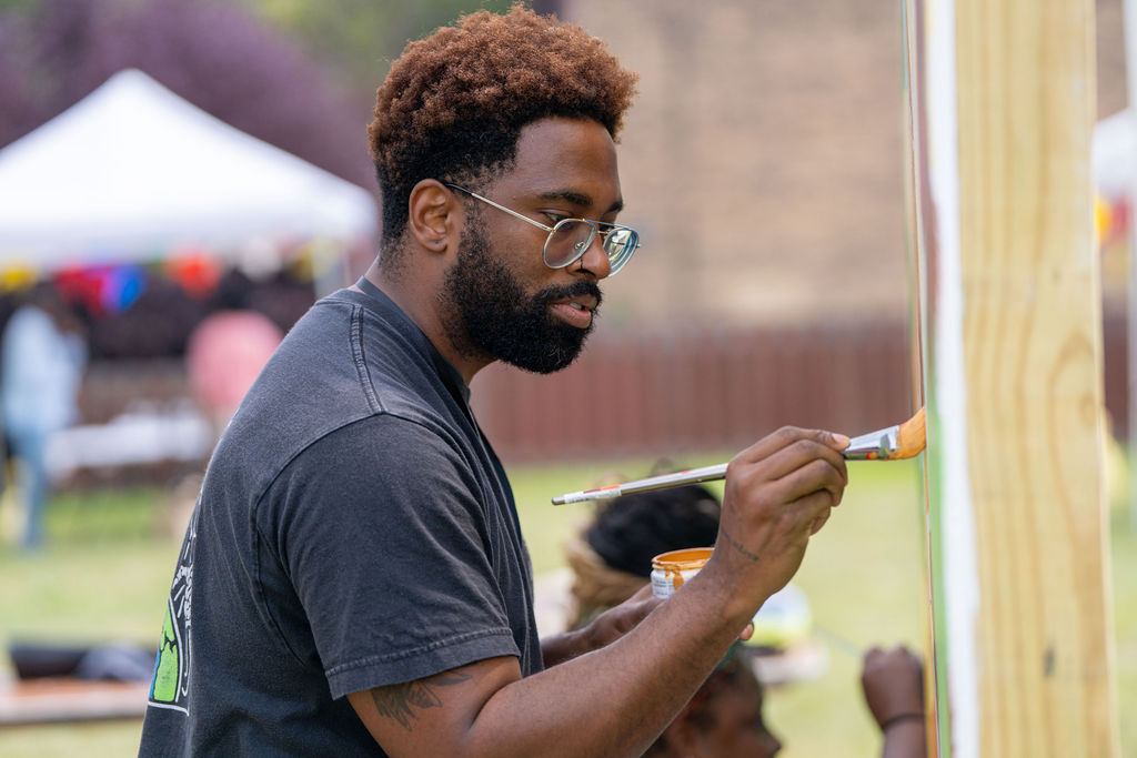 An artist paints an orange section of a wood-framed mural panel outdoors at a community event.