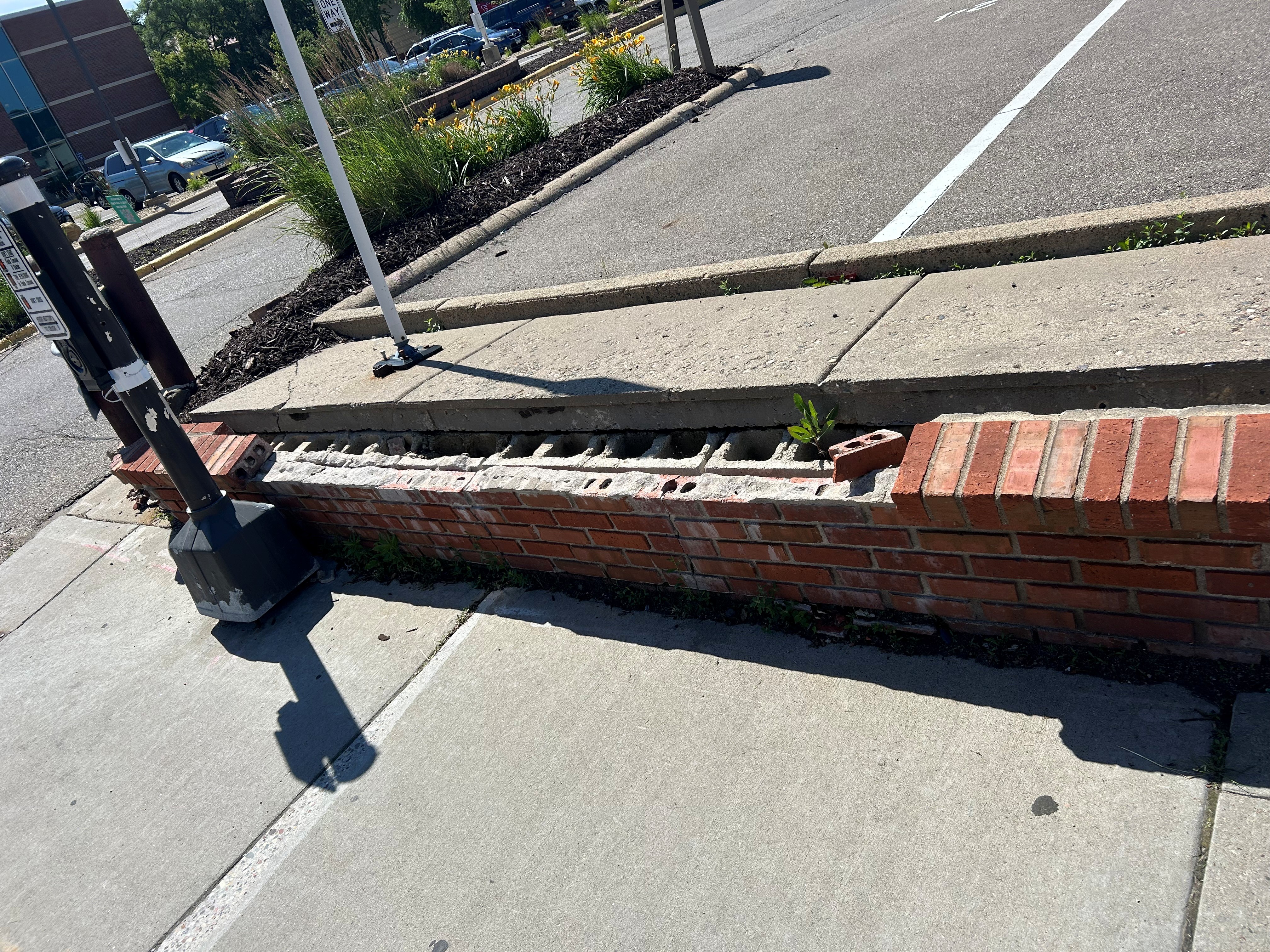 A damaged section of a low red-brick retaining wall along a sidewalk, with the top course missing and the concrete cap partially collapsed.