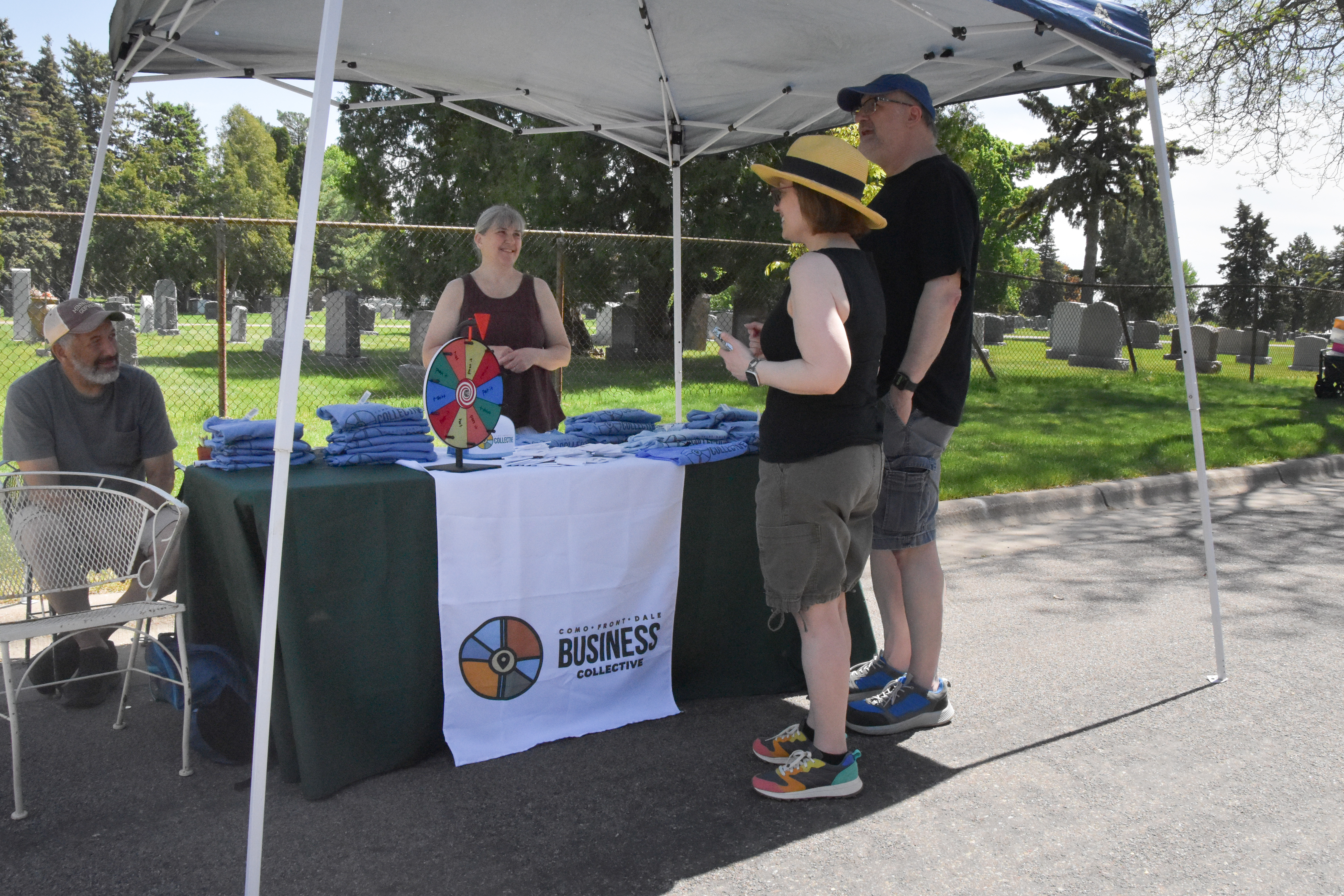 A Como Front Dale Business Collective outreach tent set up alongside Oakland Cemetery, with a staff member, T-shirts, a prize wheel and two visitors.