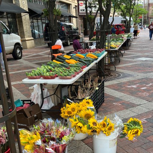An outdoor farmers market on West 7th Street with tables of fresh produce and buckets of sunflower bouquets in the foreground.