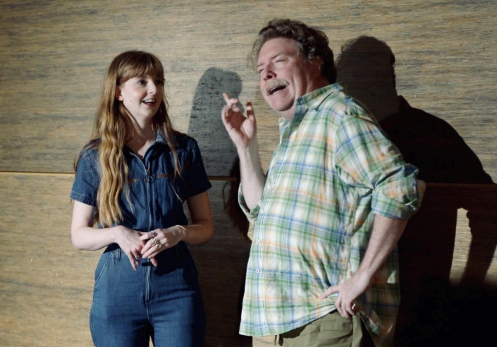 Two performers on a stage in front of a wood-paneled backdrop, both mid-performance.