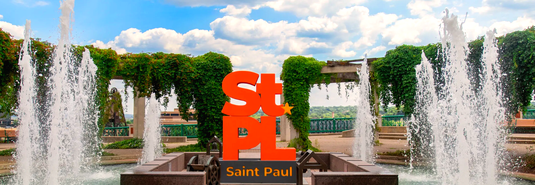The orange "StPL Saint Paul" sculpture with a orange star is composited into a scene at Upper Landing / Kellogg Park, framed by tall arcing water jets from a fountain on either side. An ivy-covered wooden pergola, park benches, a plaza and a treeline overlooking the Mississippi River bluff extend behind, under a blue sky with scattered clouds.