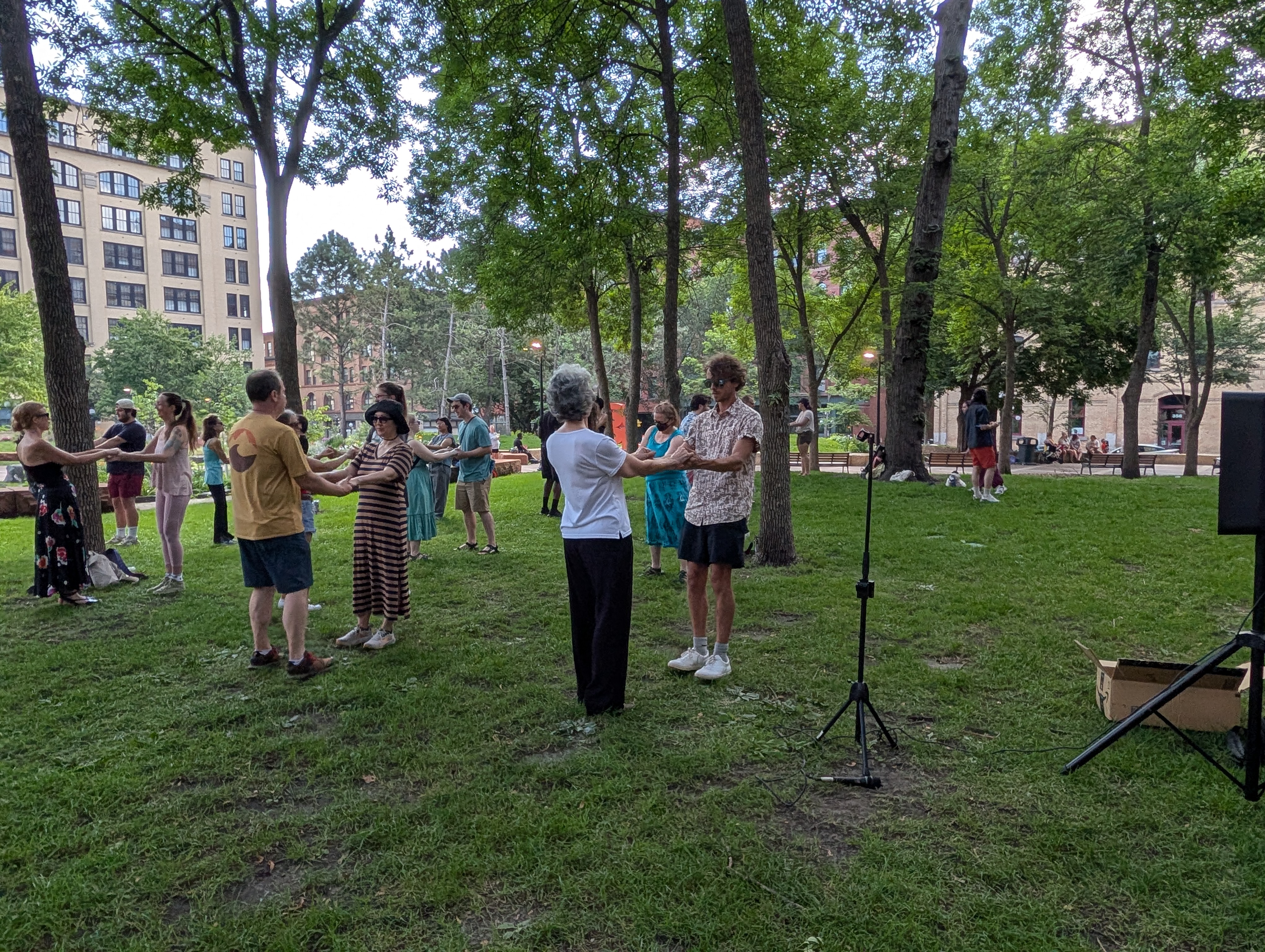 Attendees practice ballroom dance in pairs at Mears Park, with historic downtown Saint Paul buildings visible through the trees.