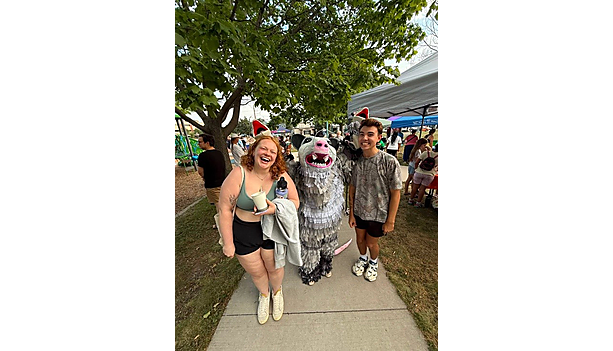 Two attendees pose with a fringed opossum mascot at an outdoor community event on a sunny sidewalk.