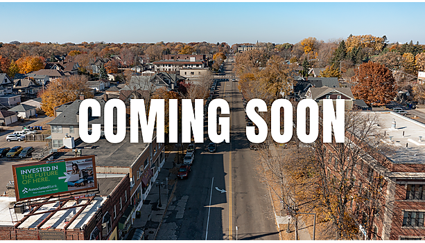 Aerial "Coming Soon" graphic over an autumn view down a Saint Paul commercial corridor with brick storefronts and a rooftop Associated Bank billboard.