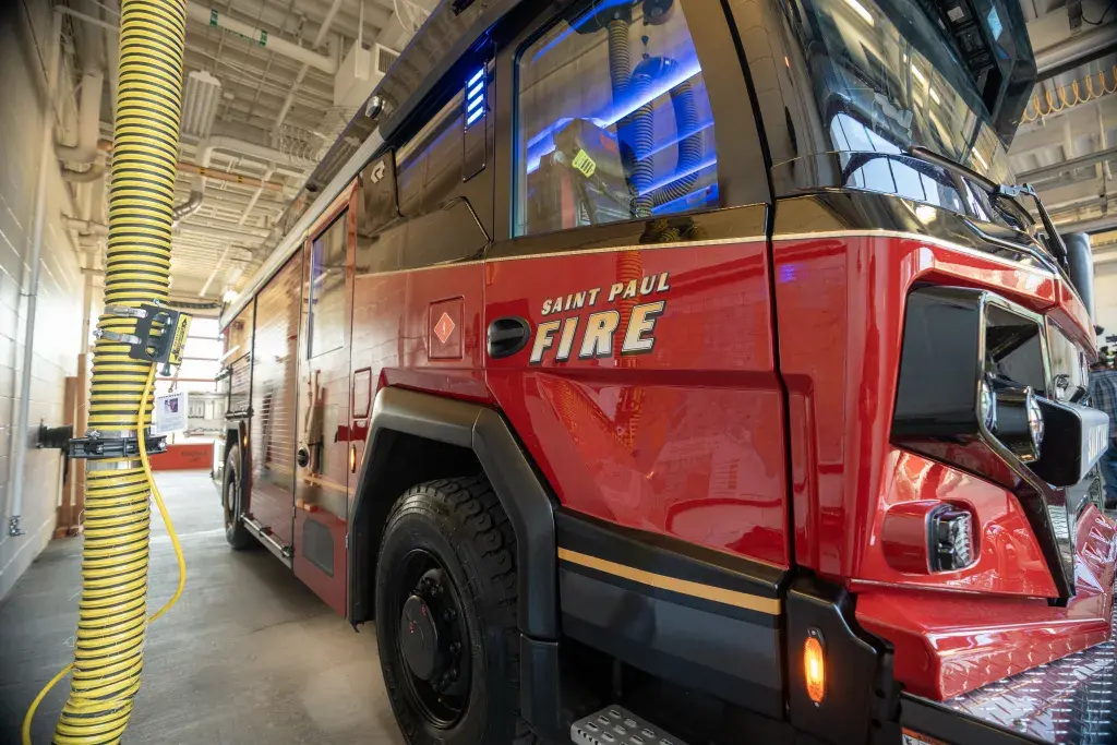 Red Saint Paul Fire truck parked inside a fire station, connected to a large yellow exhaust hose hanging from the ceiling.
