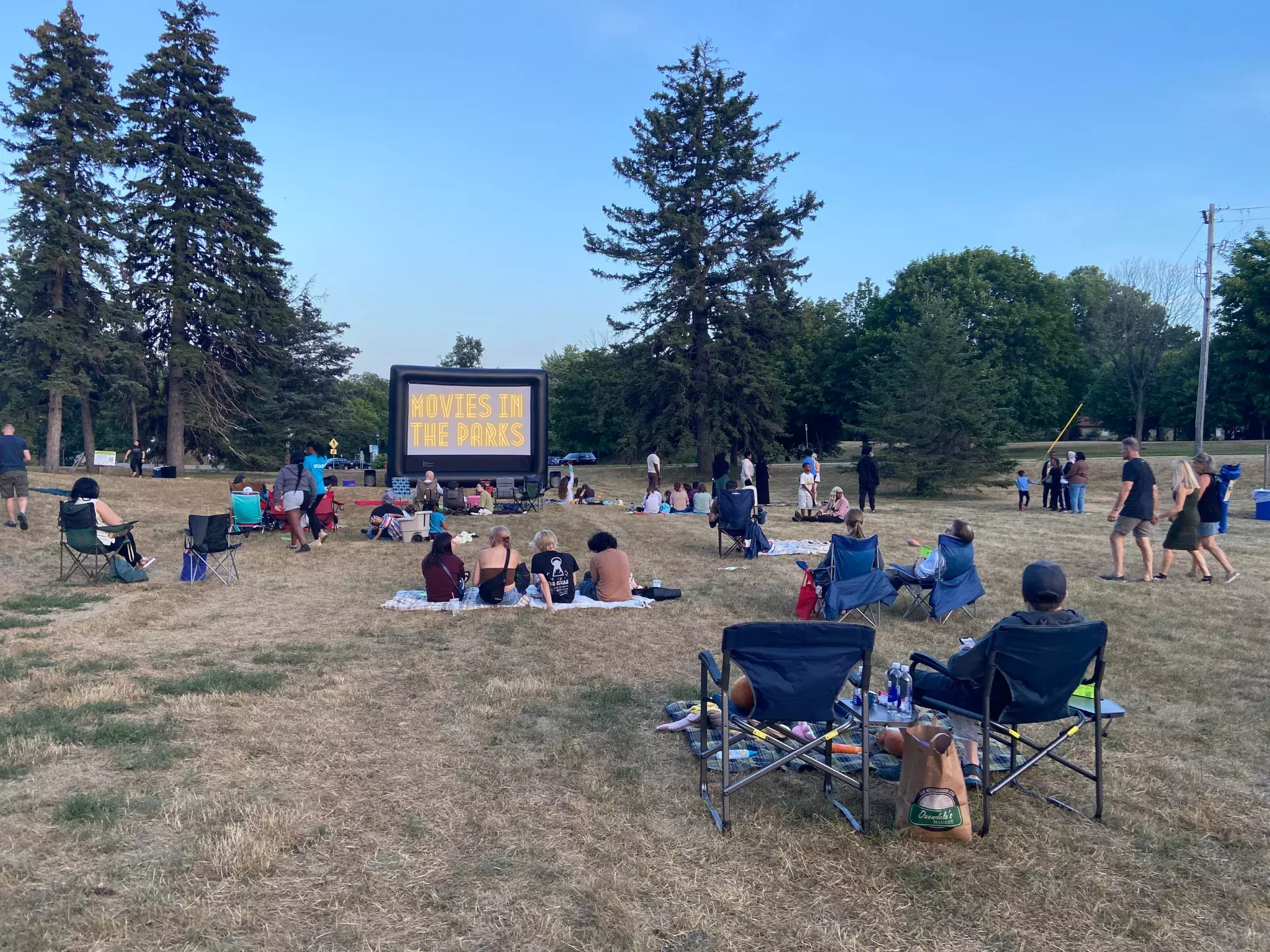 People gathered on a grassy lawn in front of an outdoor movie screen that reads "Movies in the Parks".