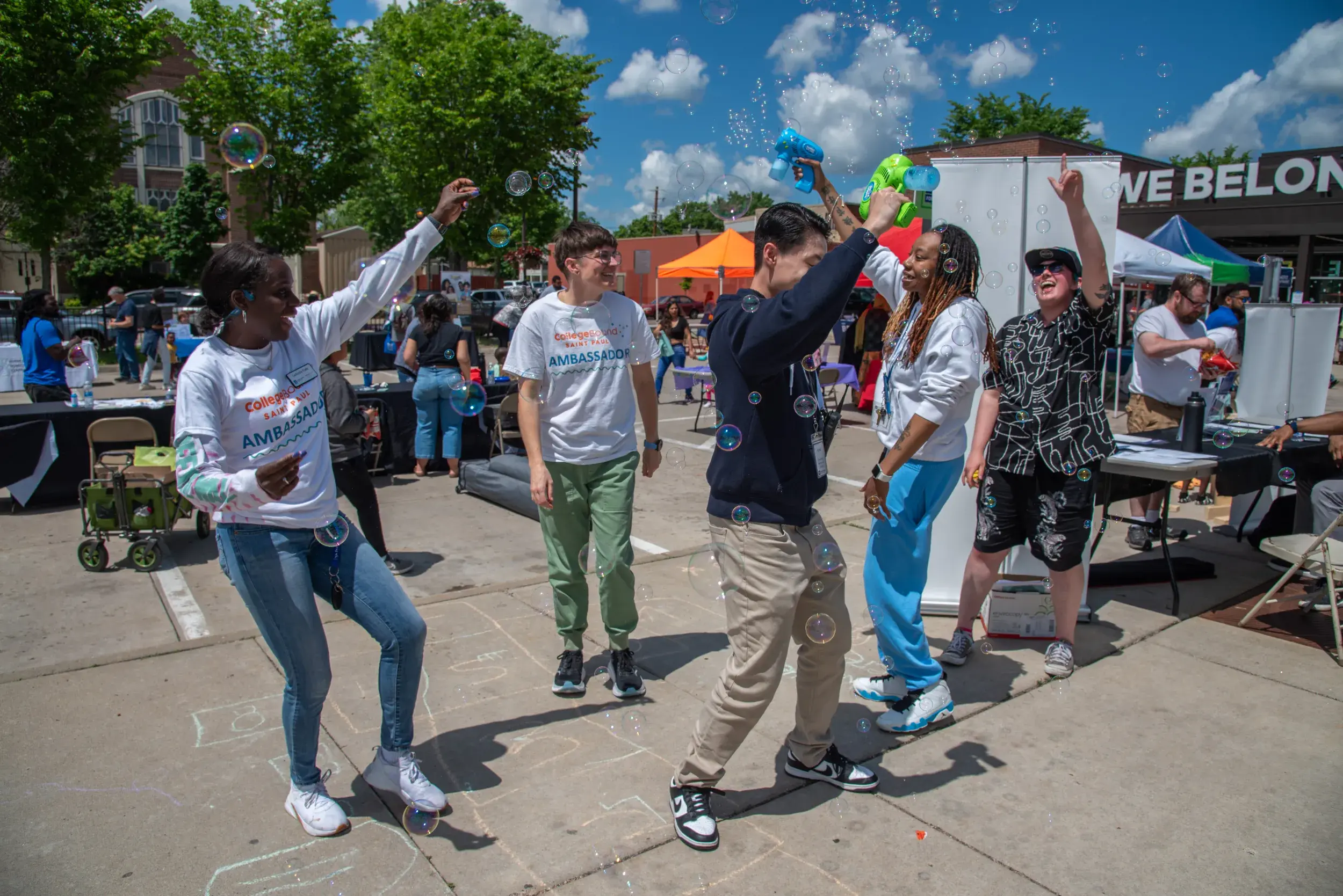 Outside a community center, people dance joyfully among a shower of iridescent bubbles on a beautiful summer day.