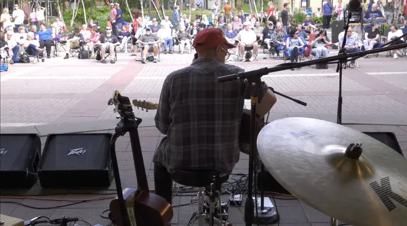 Photo of a man playing a guitar on an outdoor stage, facing an audience of people, sitting in chairs or walking around.