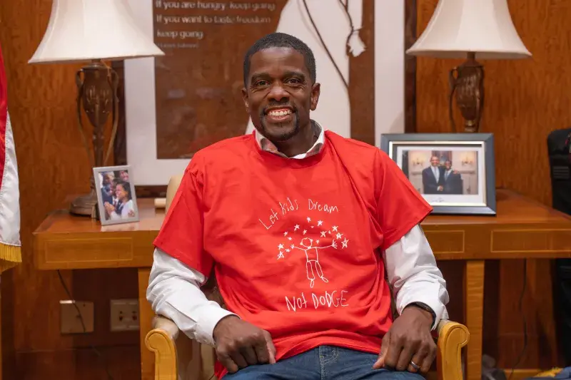 A man in a formal office models a tee-shirt that says "Let Kids Dream, Not Dodge"