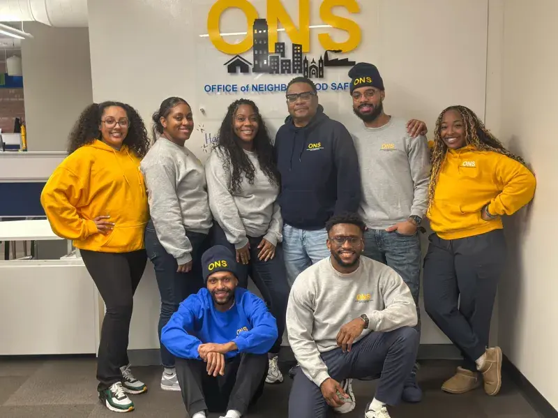 Seven smiling people pose in front of a sign that says ONS: Office of Neighborhood Safety.