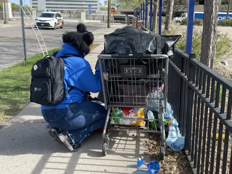 A woman kneels on the sidewalk next to a grocery cart that’s full of someone’s belongings.