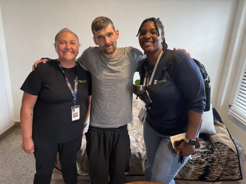 A man and two women pose, smiling, for a picture. The man has his arms around the two women's shoulders.