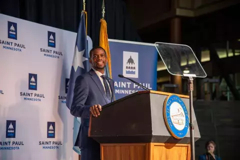 Mayor Carter speaks at podium with the City of Saint Paul seal on the front. He's in a large atrium with flags and an official City backdrop behind him.
