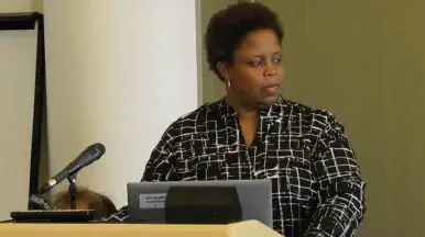 A woman in a black-and-white patterned top stands at a podium with a microphone and laptop, looking to the side during a presentation.